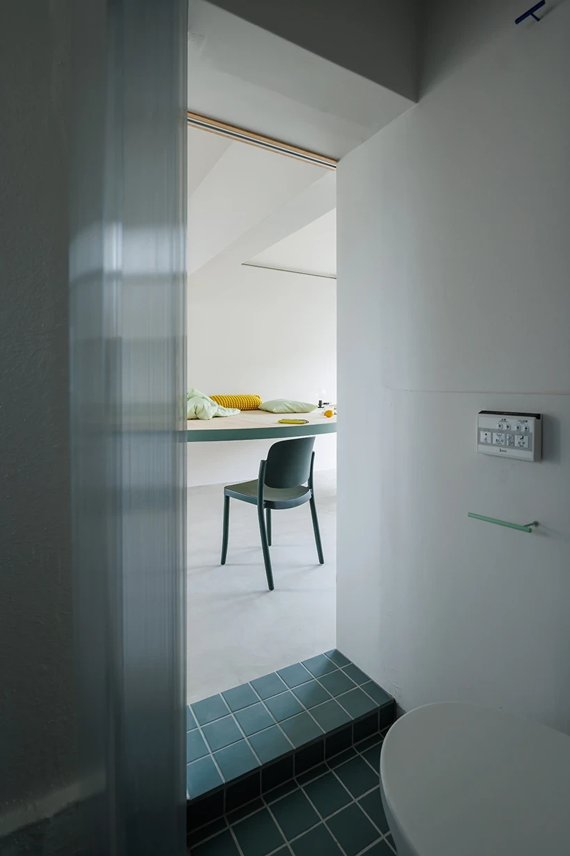 A Large Semicircle That Holds Everyday Life by Tato Architects, photographed by Yohei Sasakura: seen from the toilet side through a translucent corrugated screen, teal floor tiles match the table edge, a green chair and the table's underside beyond.