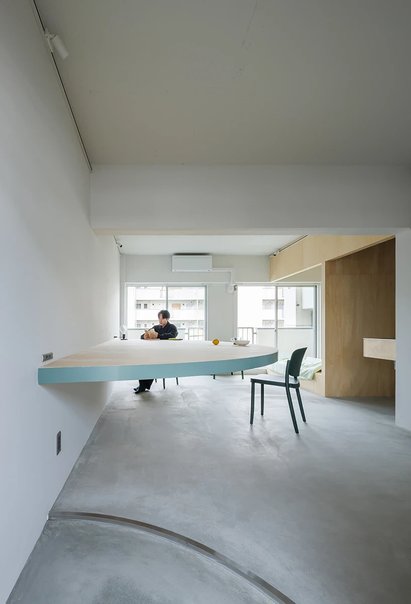 A Large Semicircle That Holds Everyday Life by Tato Architects, photographed by Yohei Sasakura: a portrait view with a man seated beneath the levitating table, green chairs distributed through the room and the plywood volume behind.