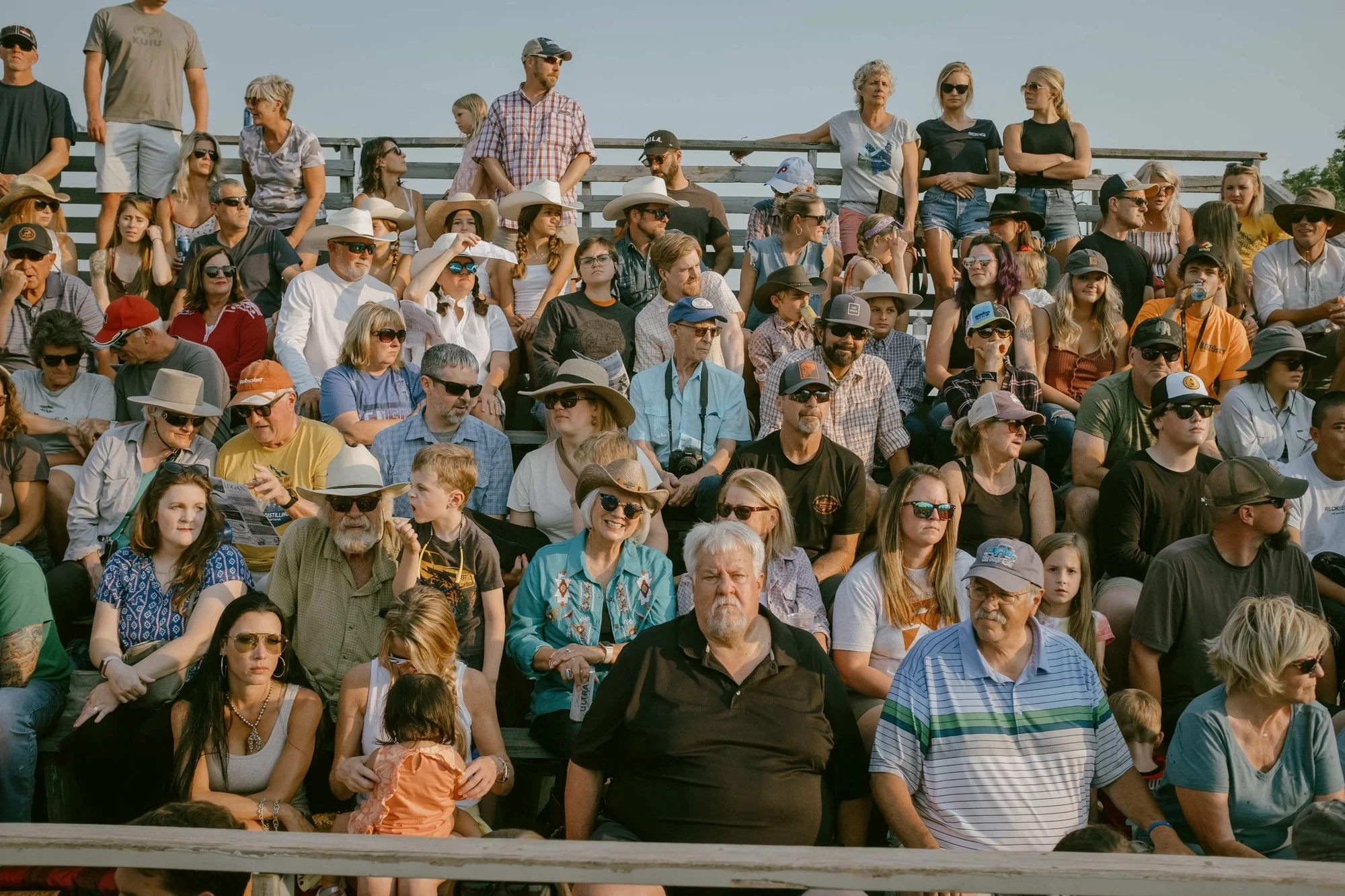 Brash by Aaron Agosto — A packed grandstand of spectators in caps, sunglasses and cowboy hats, warm evening light raking across their faces as they watch the arena