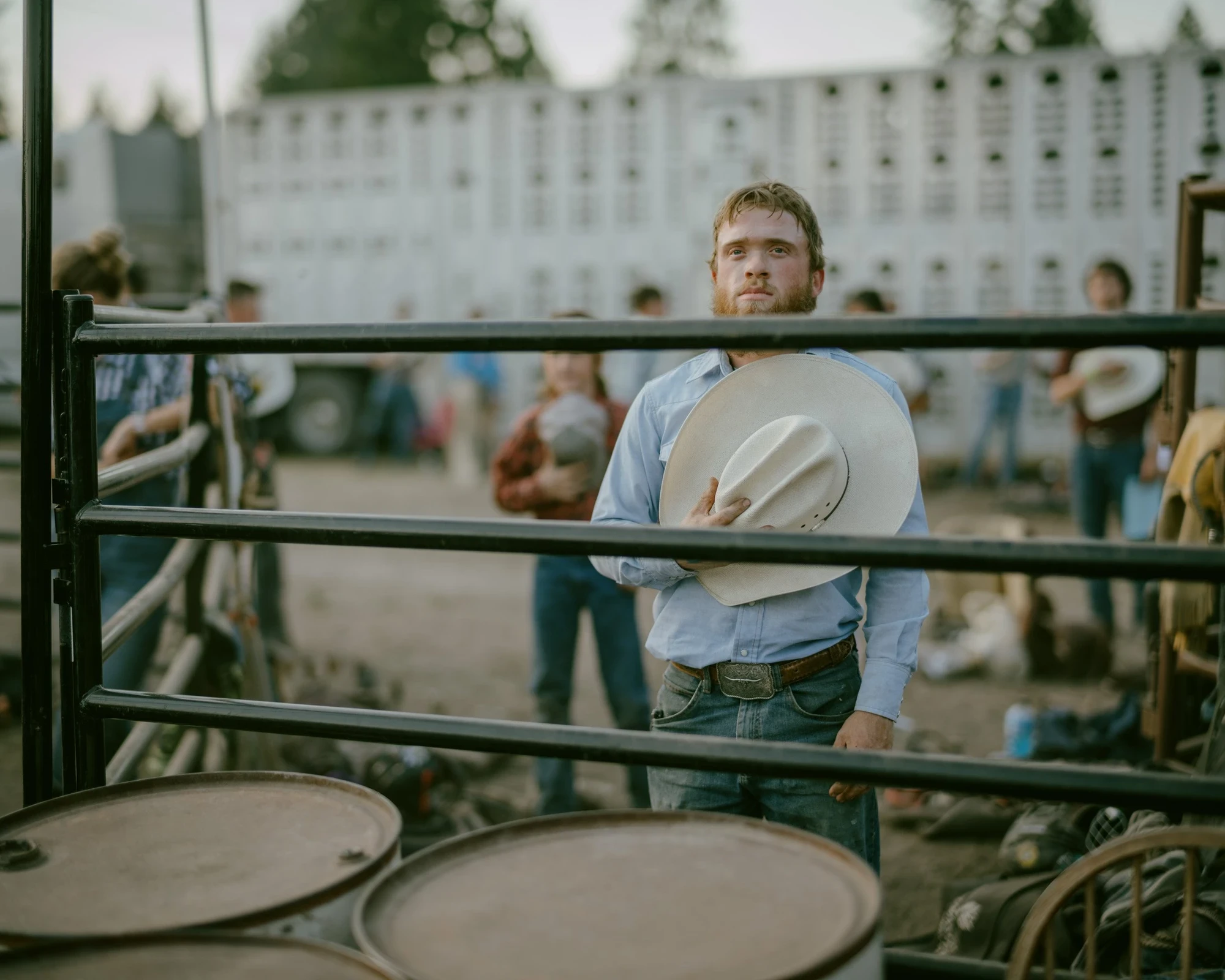 Brash by Aaron Agosto — A bearded young man in a blue shirt leans against black metal fencing holding his pale straw hat to his chest, a livestock trailer behind him