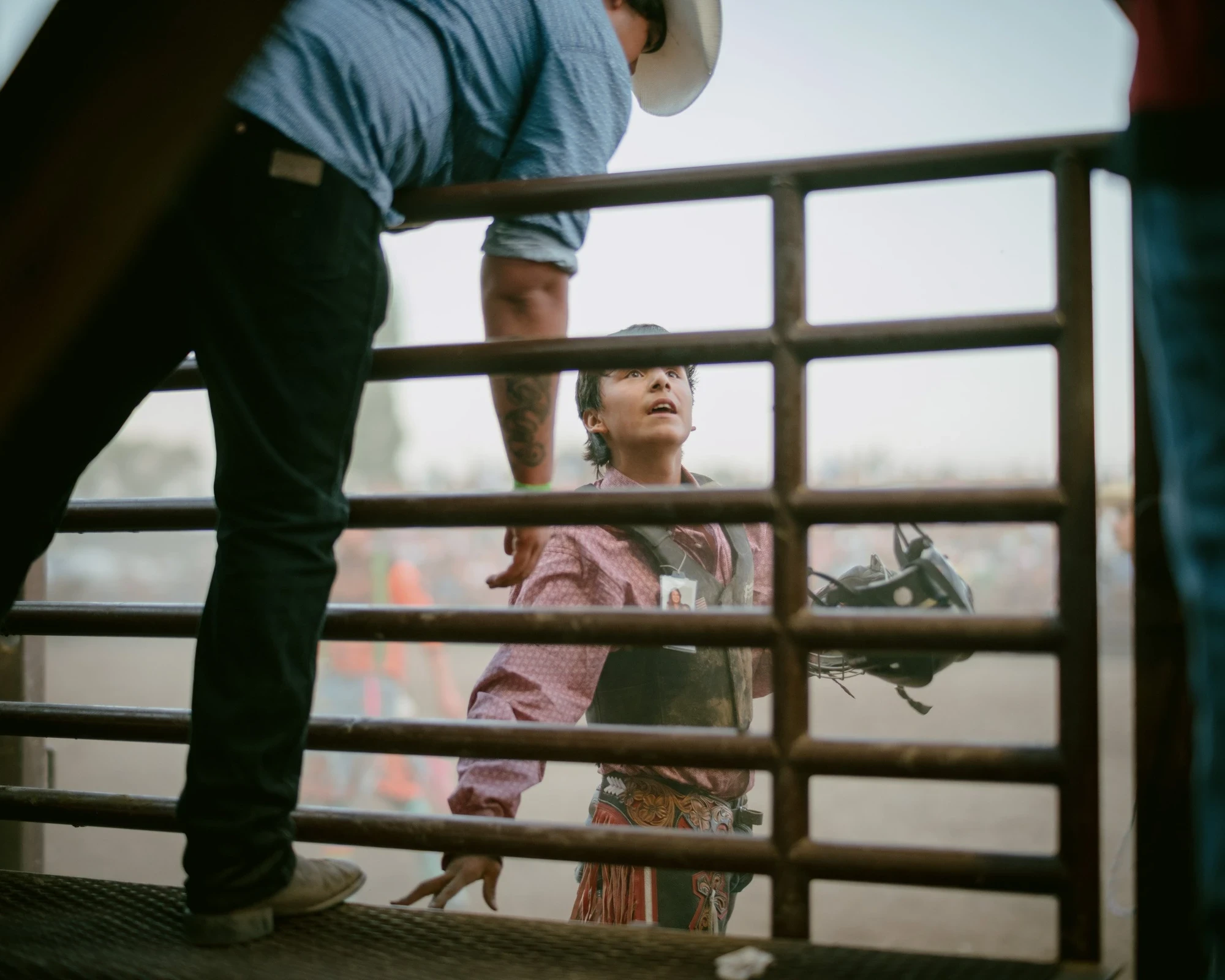 Brash by Aaron Agosto — Framed through metal chute bars, a young rider in a pink shirt and tooled chaps looks up wide-eyed while a man leans over the gate above