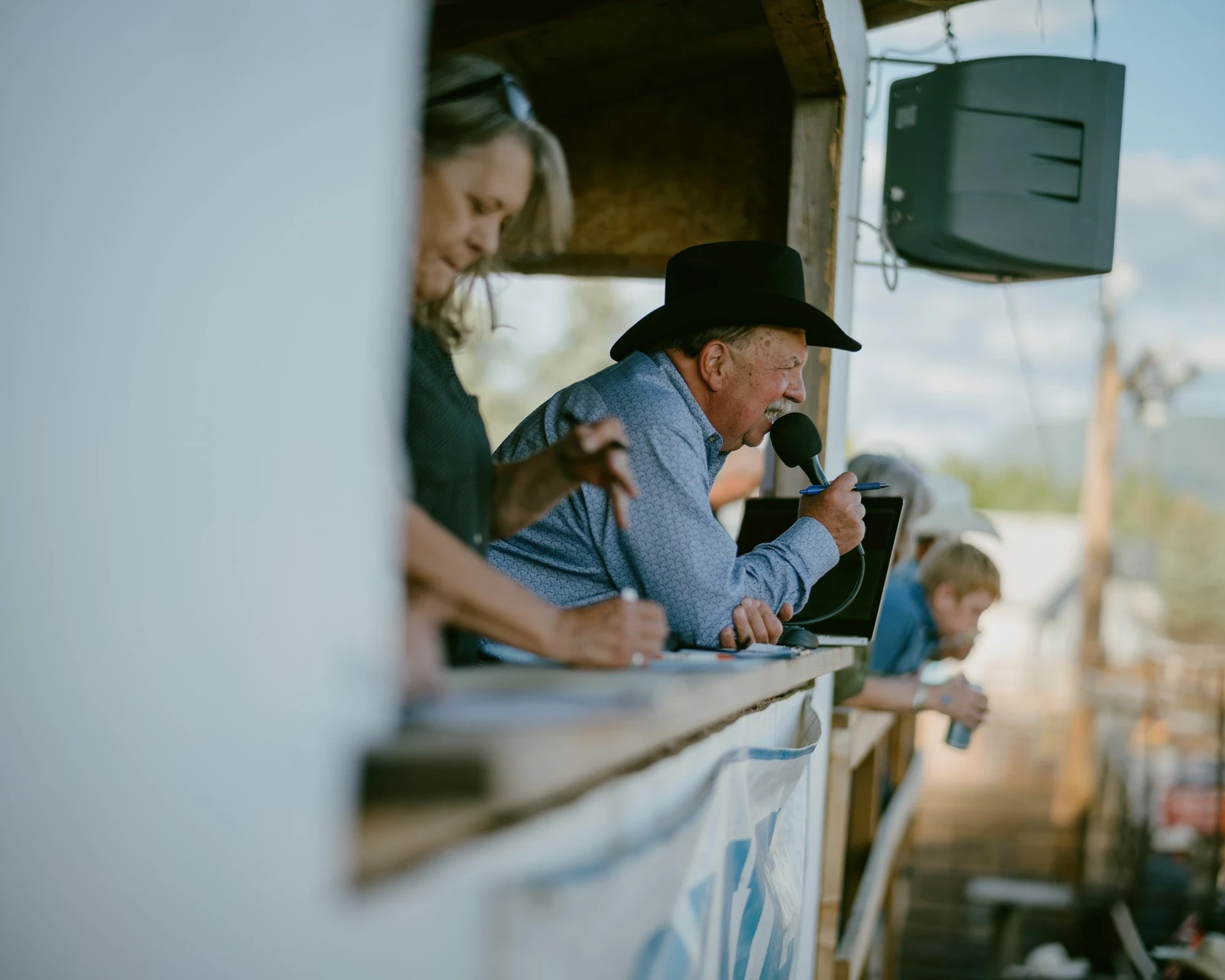 Brash by Aaron Agosto — Inside a plywood booth an announcer in a black hat speaks into a microphone beside a woman taking notes, a hanging speaker overhead