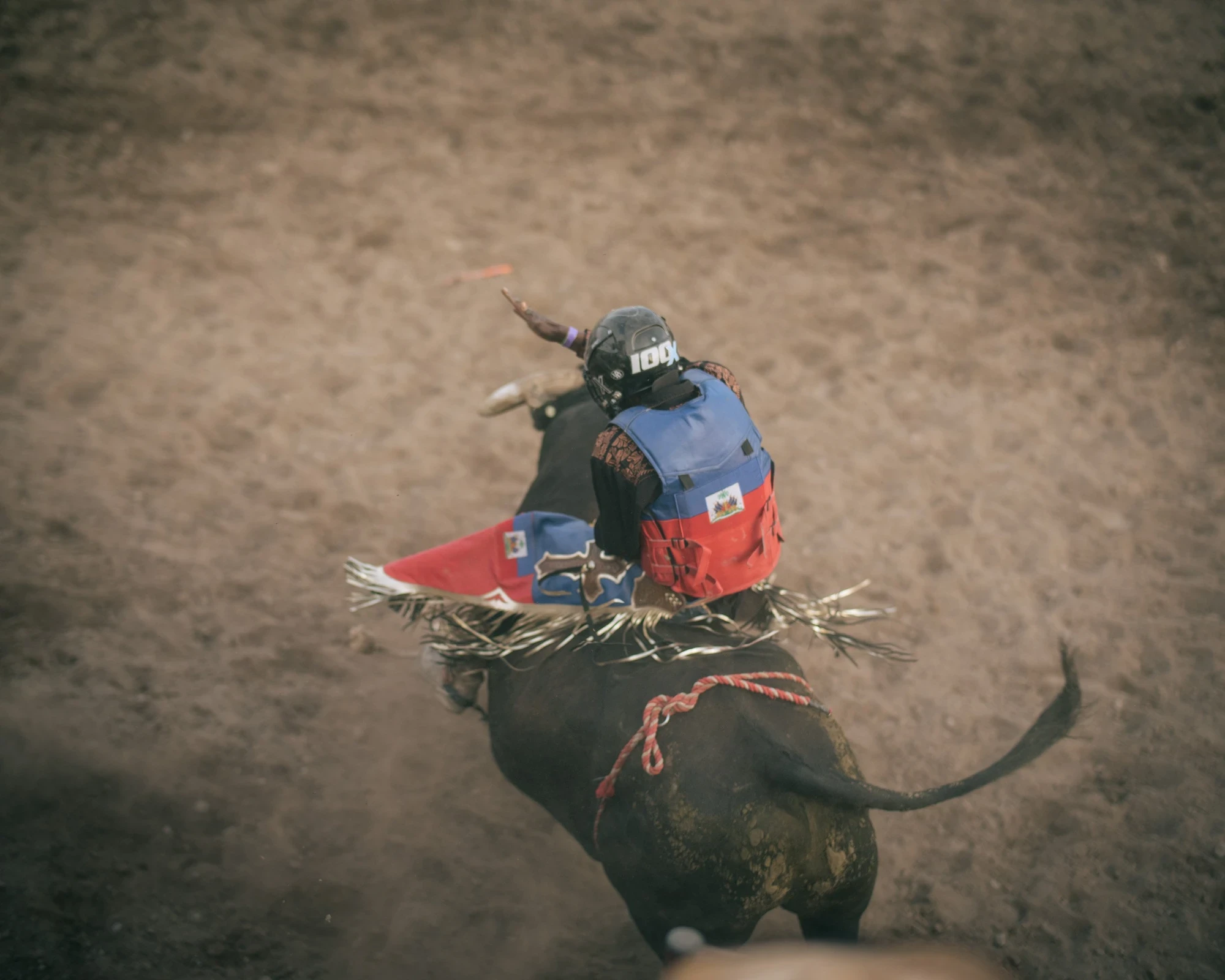 Brash by Aaron Agosto — Shot from above, a bull rider in a blue and red vest bearing the Haitian flag clings to a dark bucking bull throwing dust across the dirt