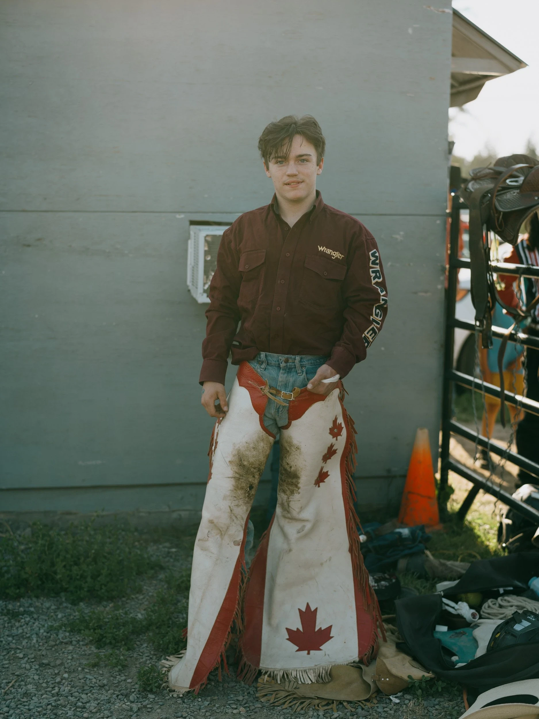 Brash by Aaron Agosto — A teenage rider stands against a gray shed wall in a maroon Wrangler shirt and mud-caked white chaps printed with a red Canadian maple leaf