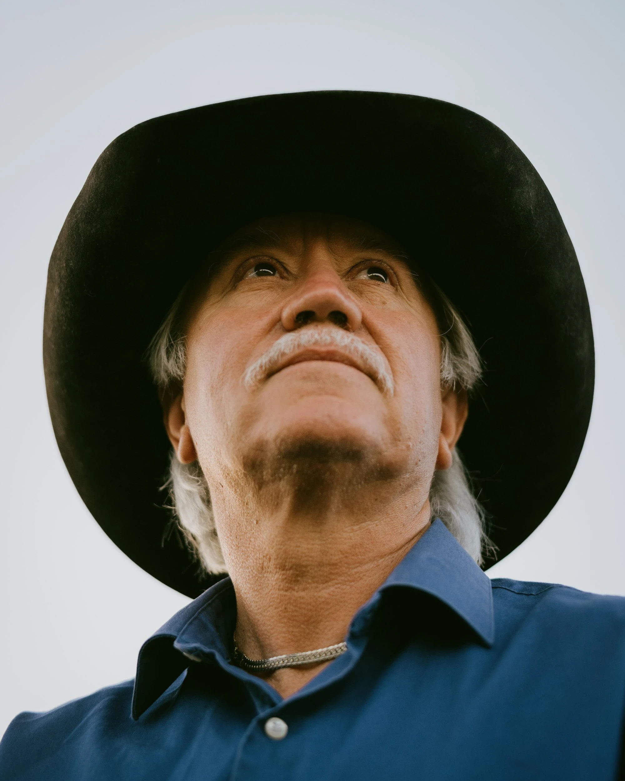 Brash by Aaron Agosto — A low-angle portrait of an older man with a white mustache in a black felt hat and blue shirt, chin lifted against flat overcast sky