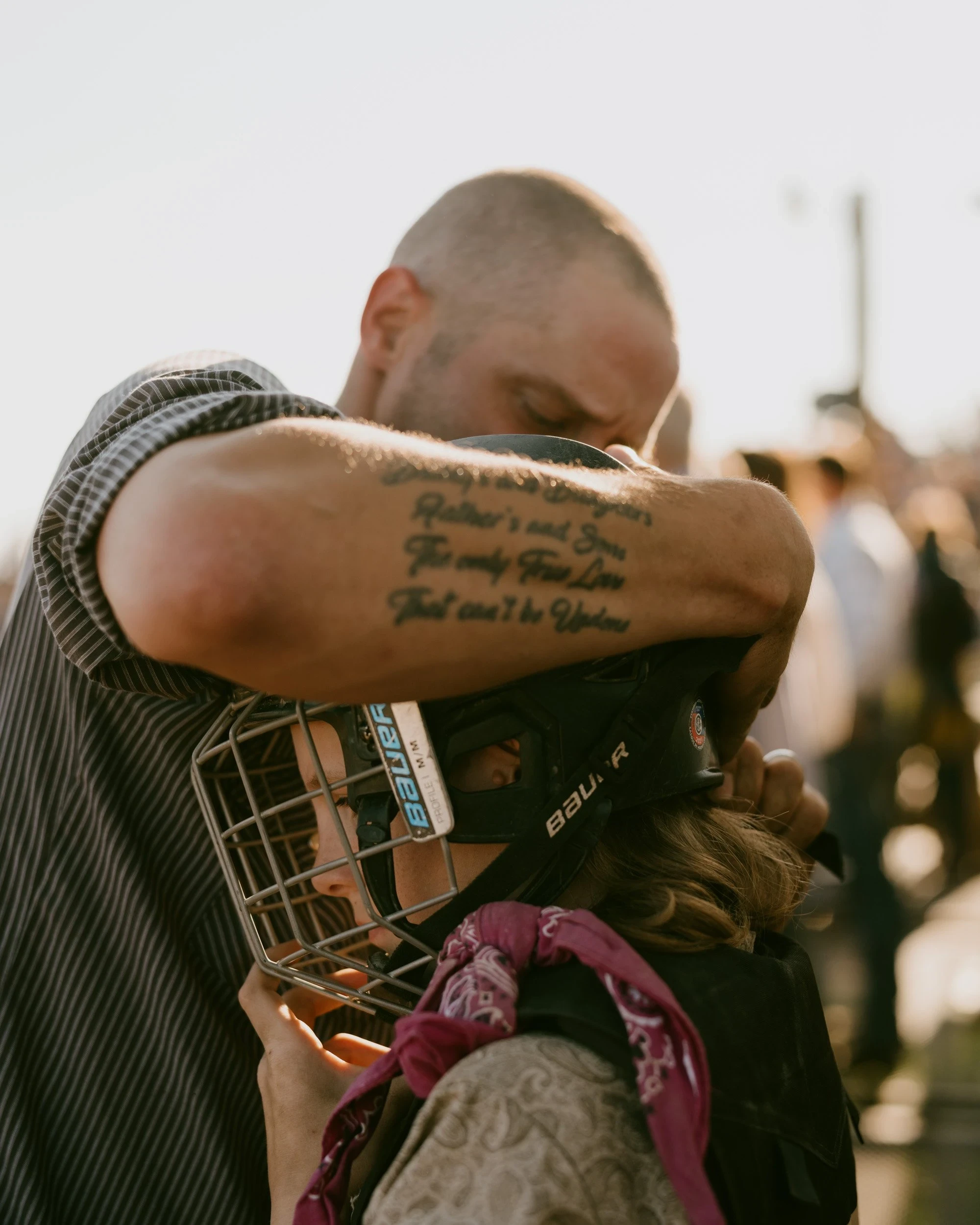 Brash by Aaron Agosto — A man presses his face into a child's caged hockey helmet, his sunlit forearm tattooed with a verse about fathers and sons