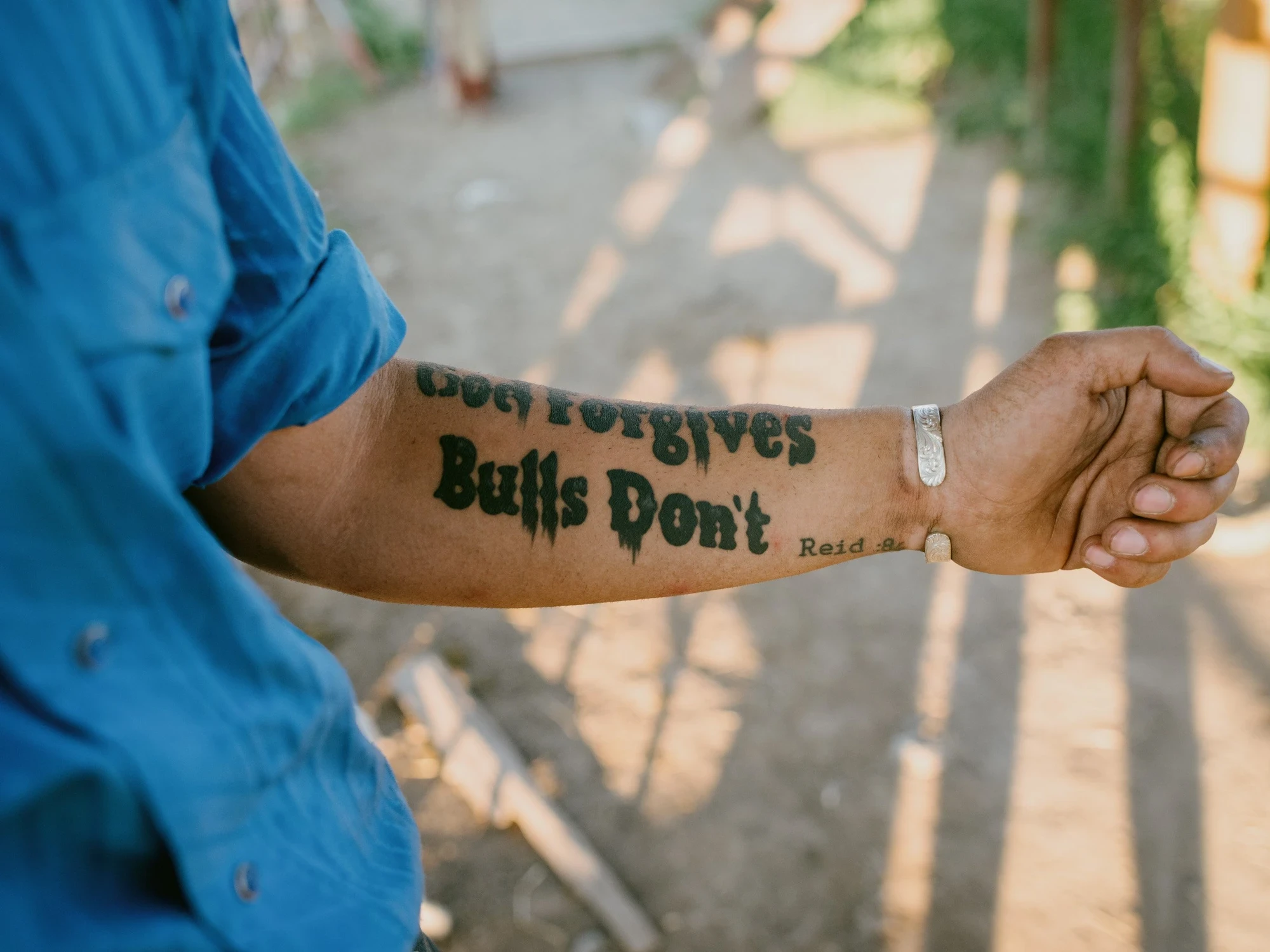 Brash by Aaron Agosto — A sunlit forearm in a blue shirt tattooed God Forgives, Bulls Don't in heavy black lettering, a silver cuff at the wrist