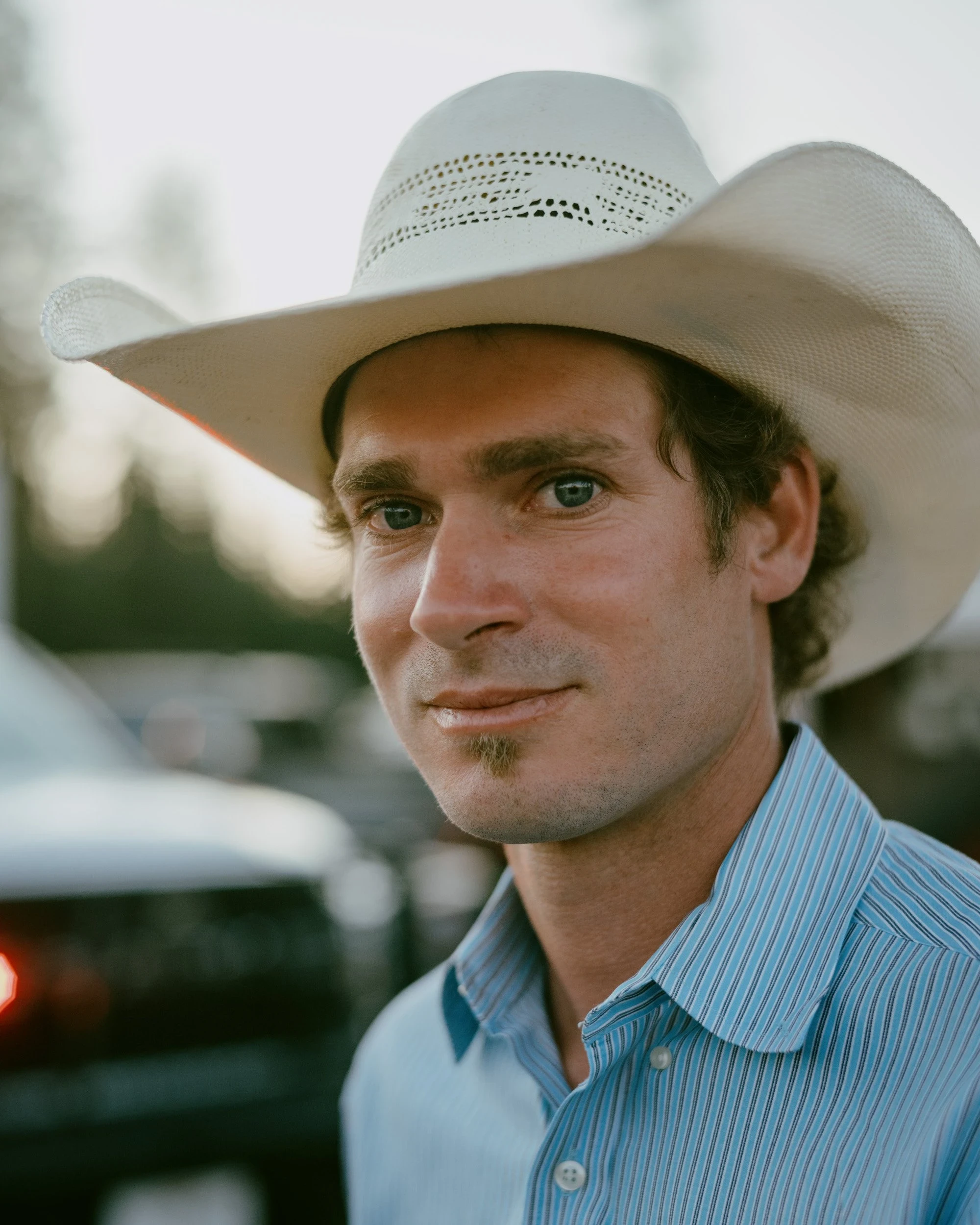 Brash by Aaron Agosto — A close portrait of a young man in a pale straw hat and blue striped shirt, faint smile, a dark truck out of focus behind him