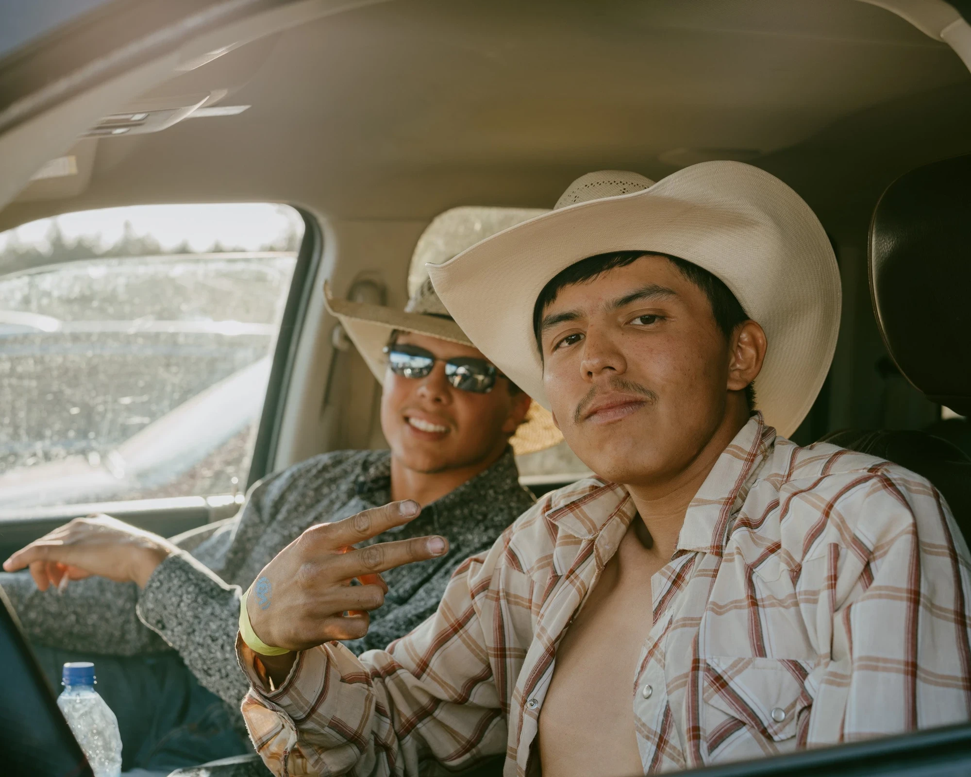 Brash by Aaron Agosto — Two young men sit in a truck cab in cowboy hats, one shirtless under an open plaid shirt throwing a peace sign, the other in sunglasses grinning