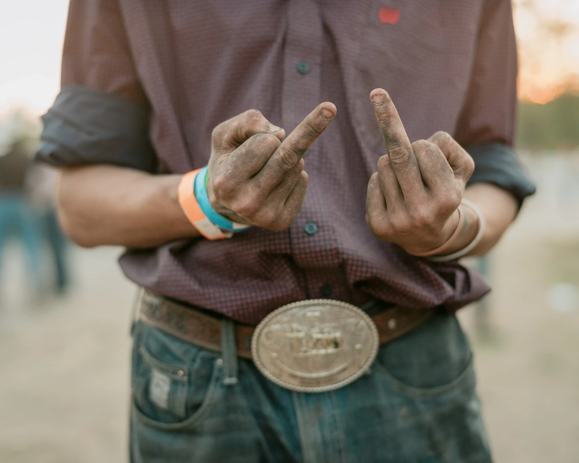 Brash by Aaron Agosto — A cropped view of dust-blackened hands raising two middle fingers over a large silver belt buckle and worn jeans at dusk