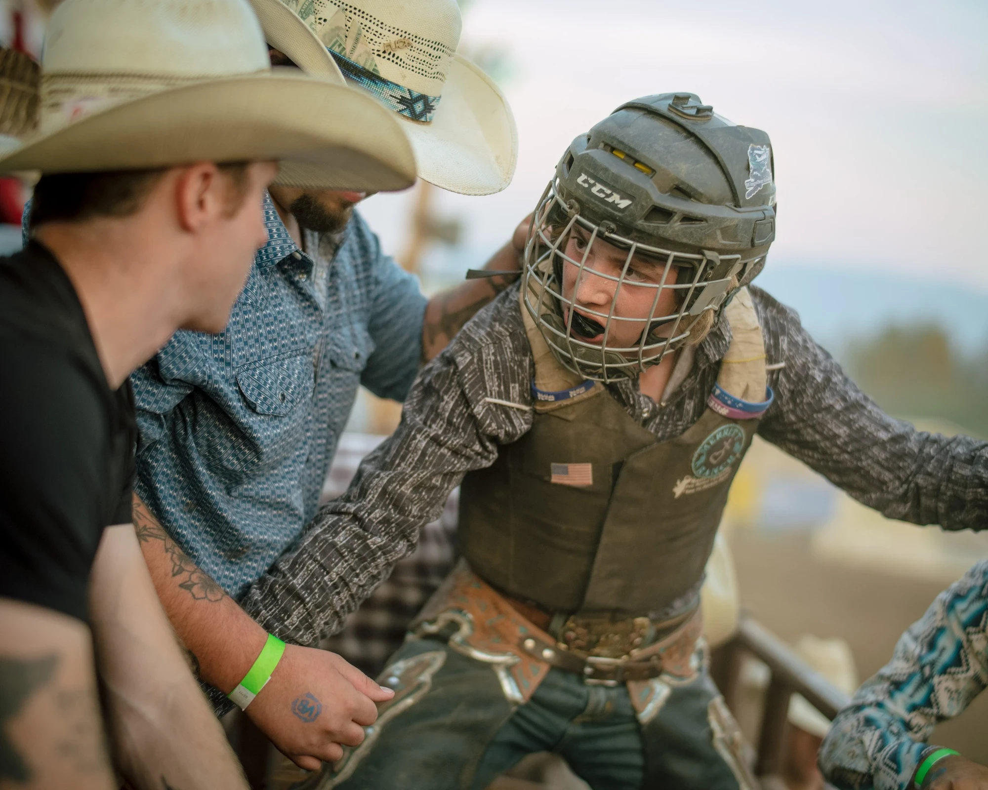 Brash by Aaron Agosto — A young rider in a caged helmet and flag-patched vest is steadied by two men, one tattooed, as evening light softens the pens behind