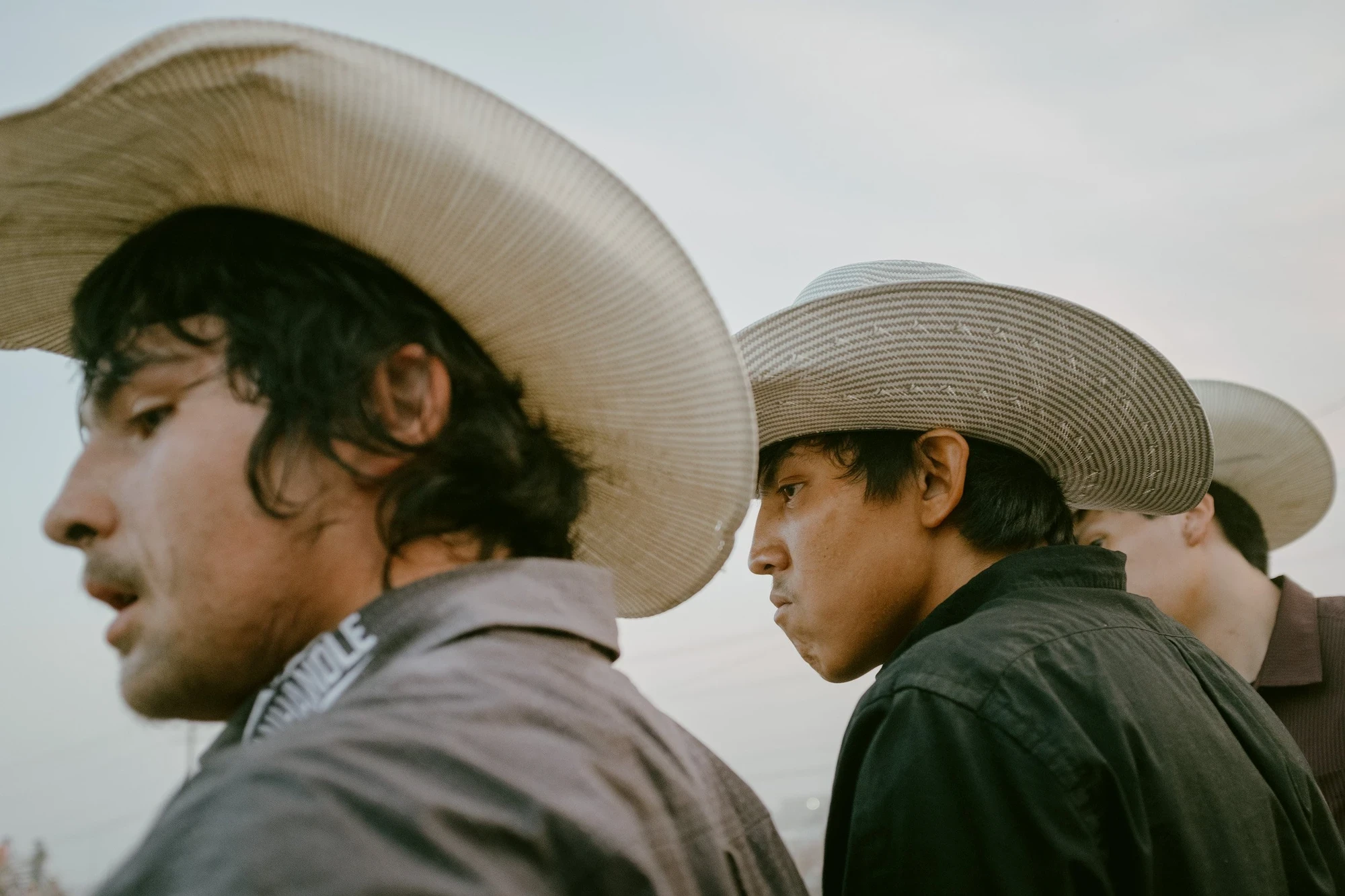 Brash by Aaron Agosto — Two young riders in straw cowboy hats seen in profile against a pale dusk sky, one hat lettered SEMINOLE, a third face just visible behind them