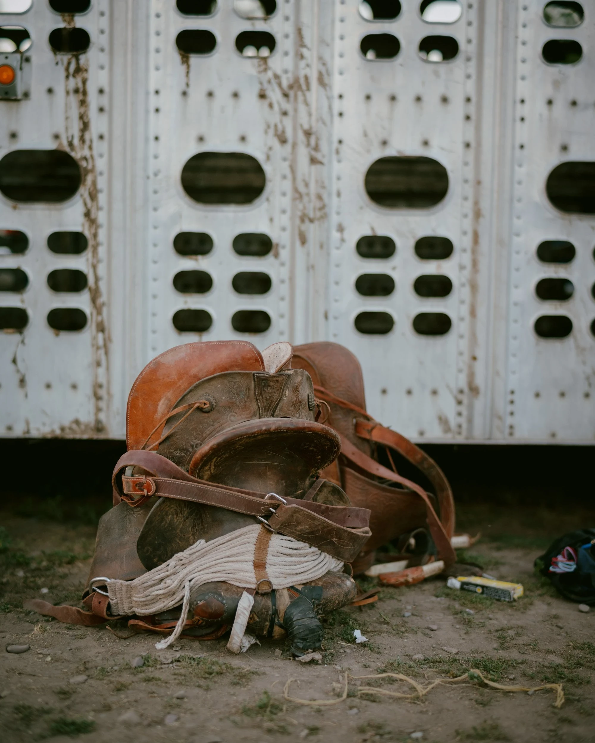 Brash by Aaron Agosto — A worn leather saddle and coiled rope slump in the dirt against the ribbed aluminum side of a livestock trailer