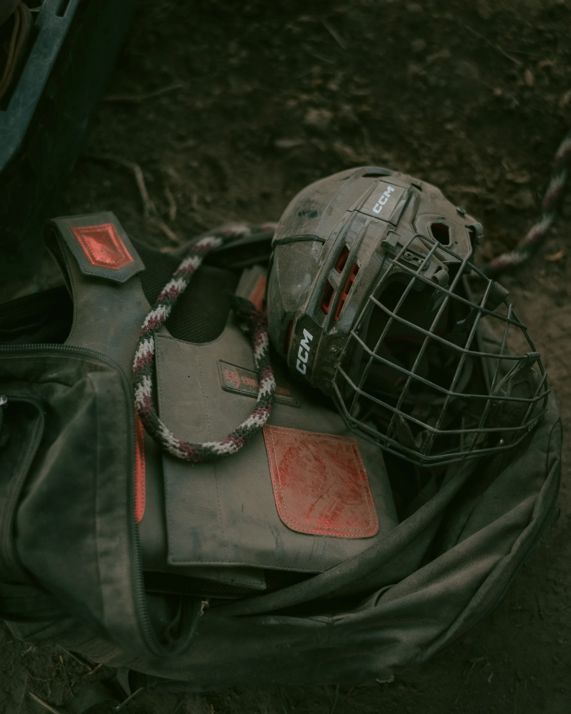 Brash by Aaron Agosto — A dust-covered CCM helmet with a wire cage rests on a protective vest inside an open gear bag on the dark ground