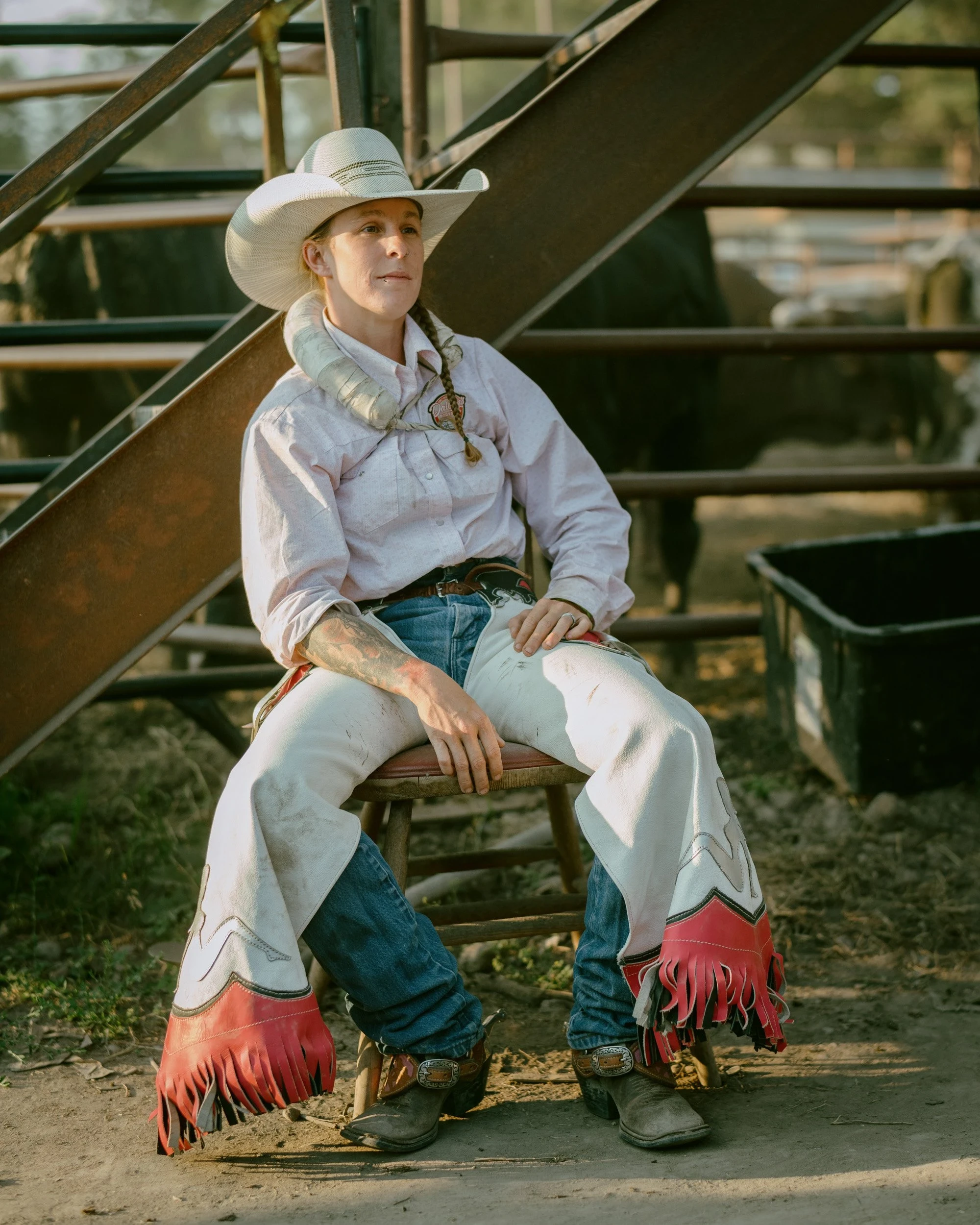 Brash by Aaron Agosto — A tattooed rider in a pale shirt and fringed white chaps sits on a wooden stool beside a metal staircase, gazing off past the pens