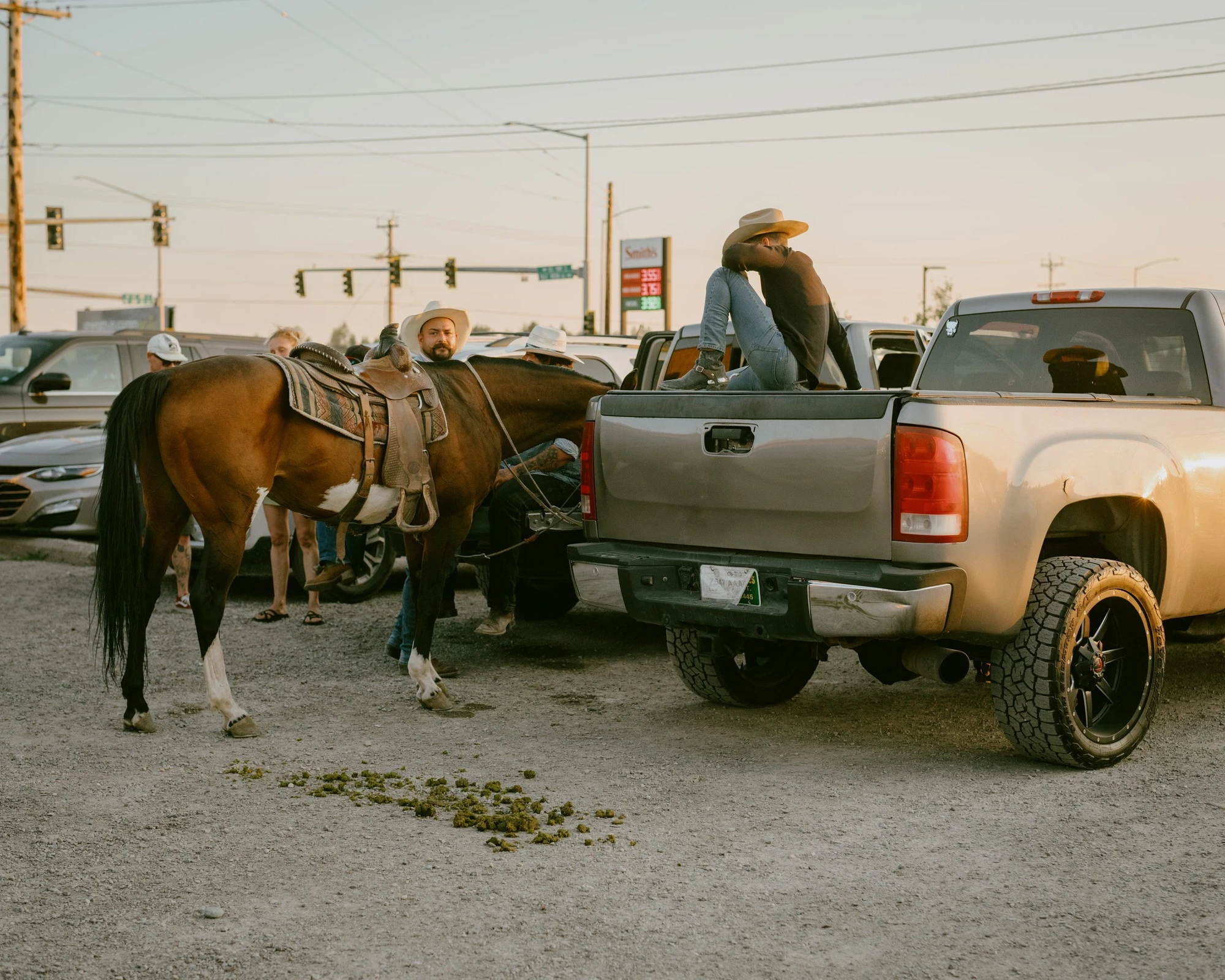 Brash by Aaron Agosto — In a gravel lot a saddled horse stands beside a silver pickup while a person in a cowboy hat rests face-down in the truck bed at dusk
