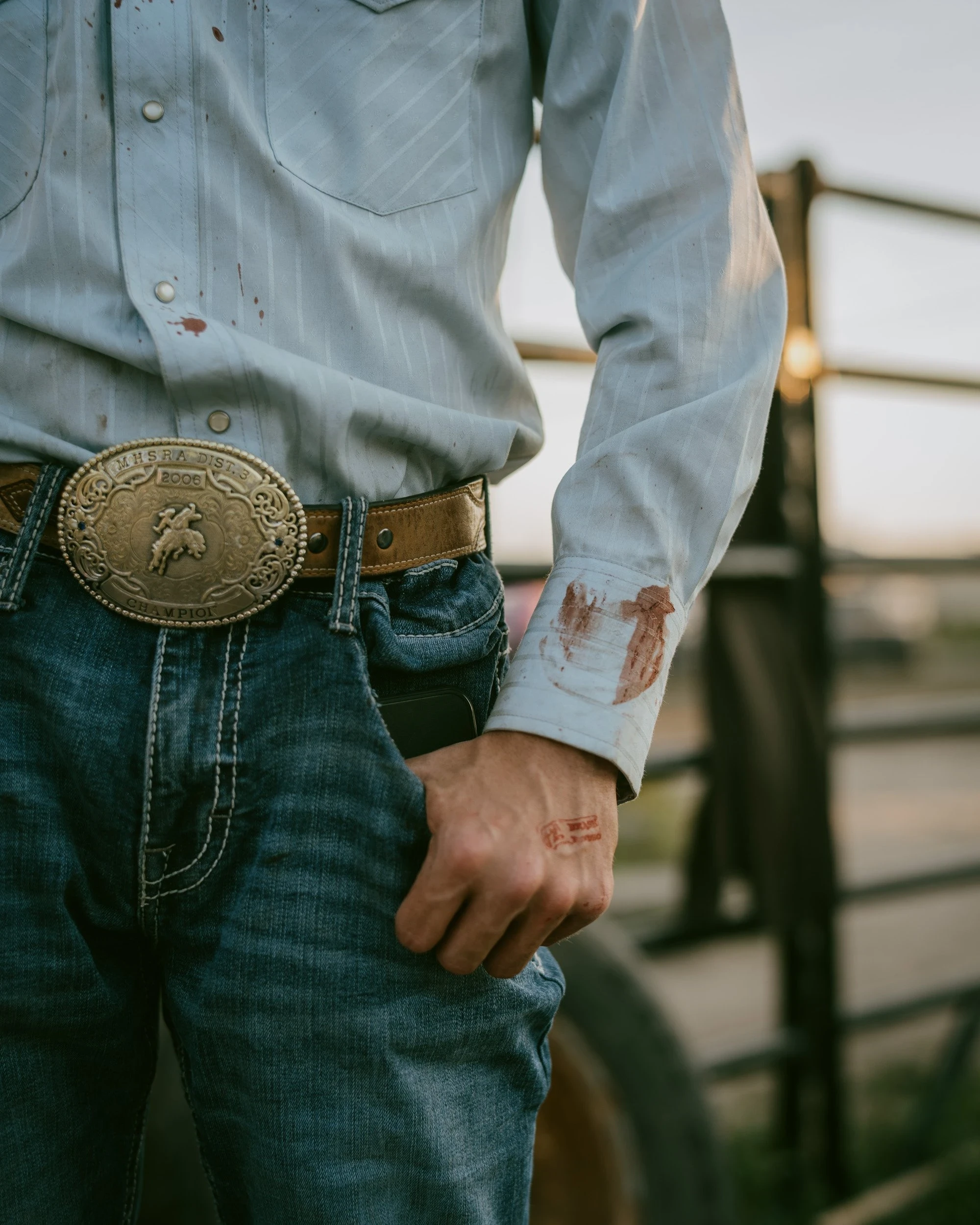 Brash by Aaron Agosto — A cropped waist-up view of a rider in a pale striped shirt with a large brass rodeo buckle, a smear of blood on the cuff and knuckle