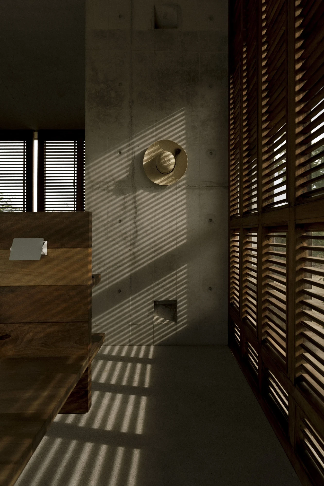Casa Viento by Aranza De Ariño, photographed by Diego Orvaños — angled louvers rake striped shadow across a board-formed concrete wall and a straw hat.