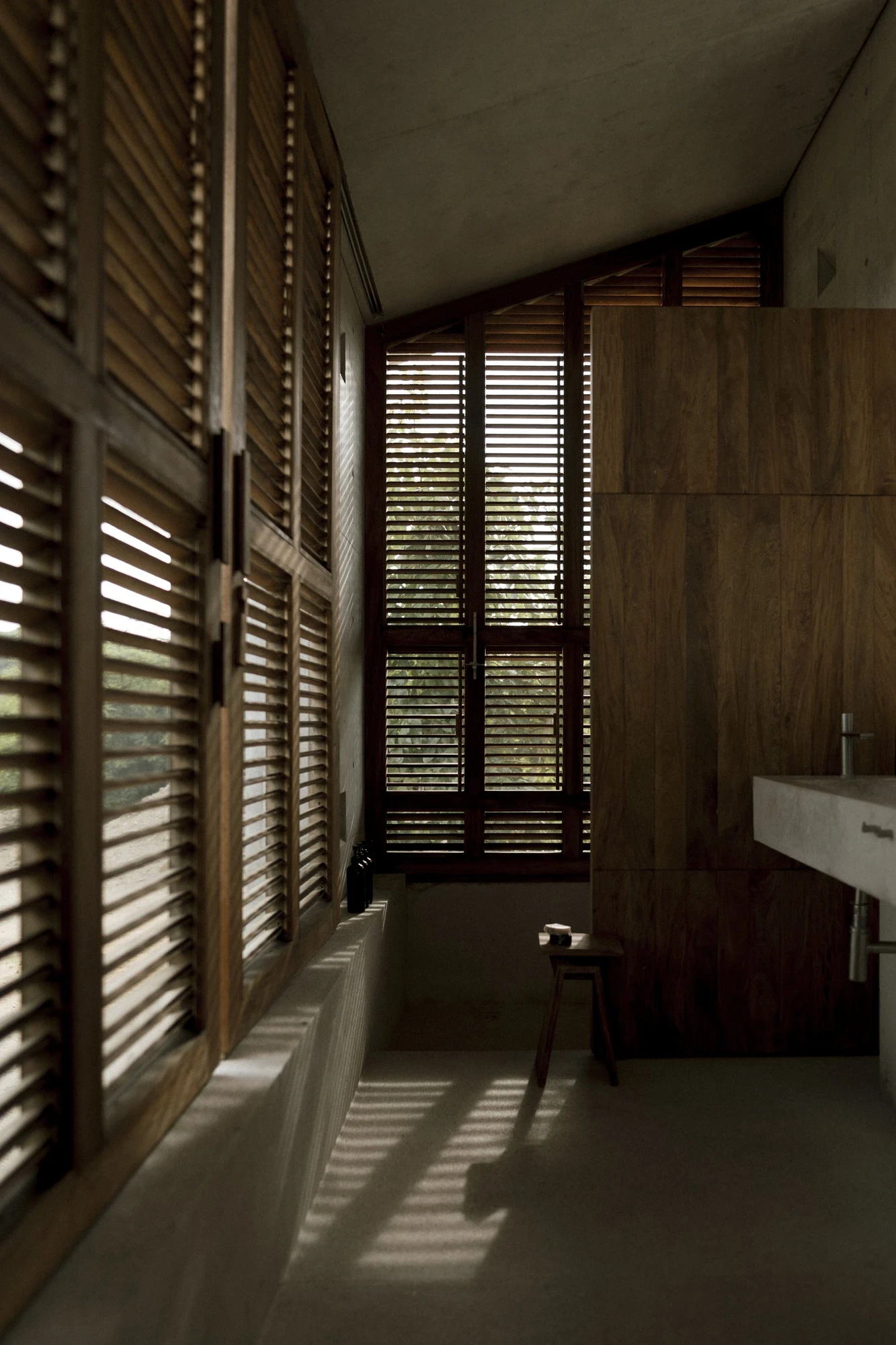 Casa Viento by Aranza De Ariño, photographed by Diego Orvaños — floor-to-ceiling teak louvers line a narrow bathroom, filtering low light onto a concrete sink.