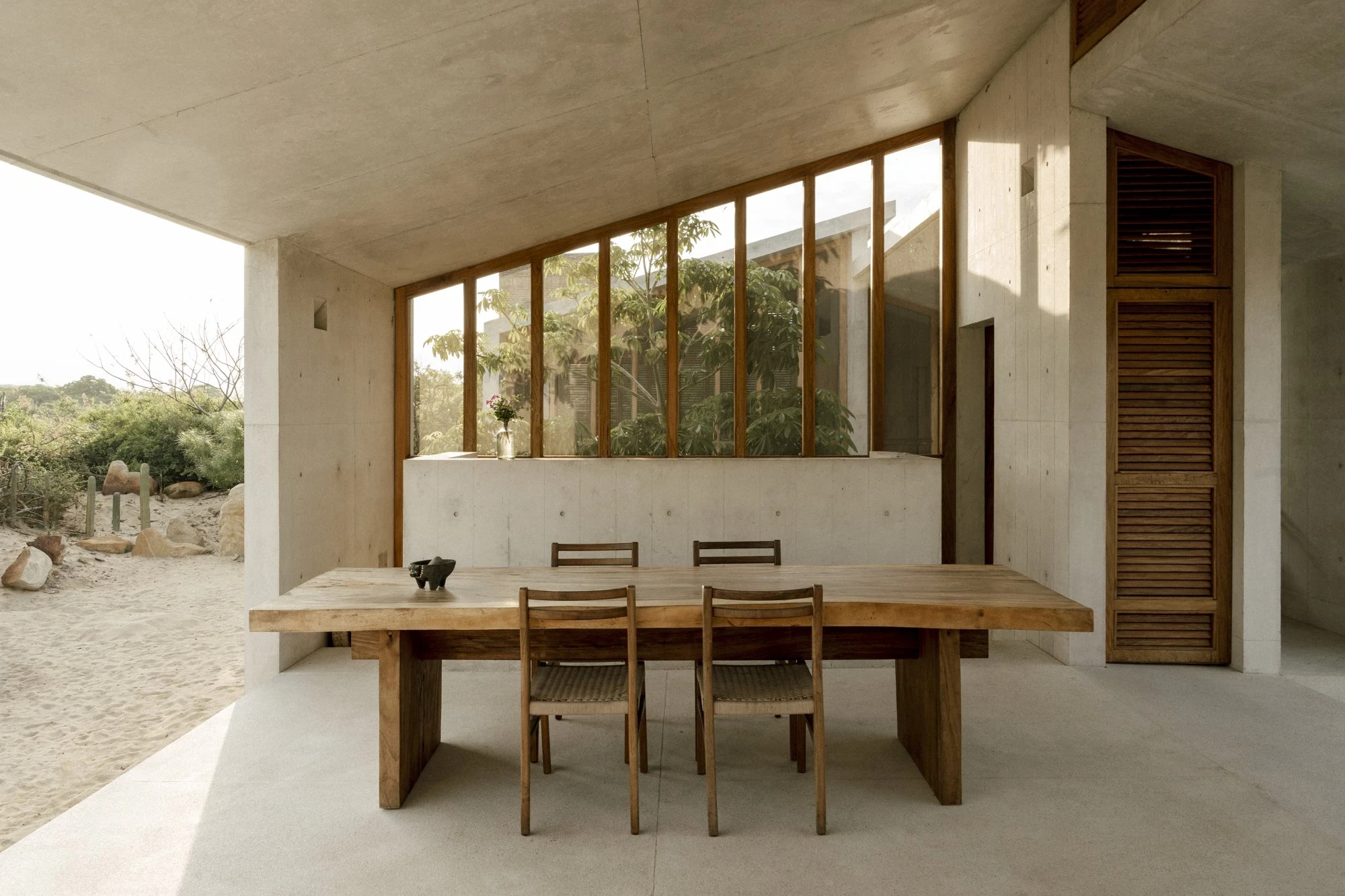 Casa Viento by Aranza De Ariño, photographed by Diego Orvaños — a live-edge timber dining table under a concrete soffit, clerestory glazing behind and sand beyond.