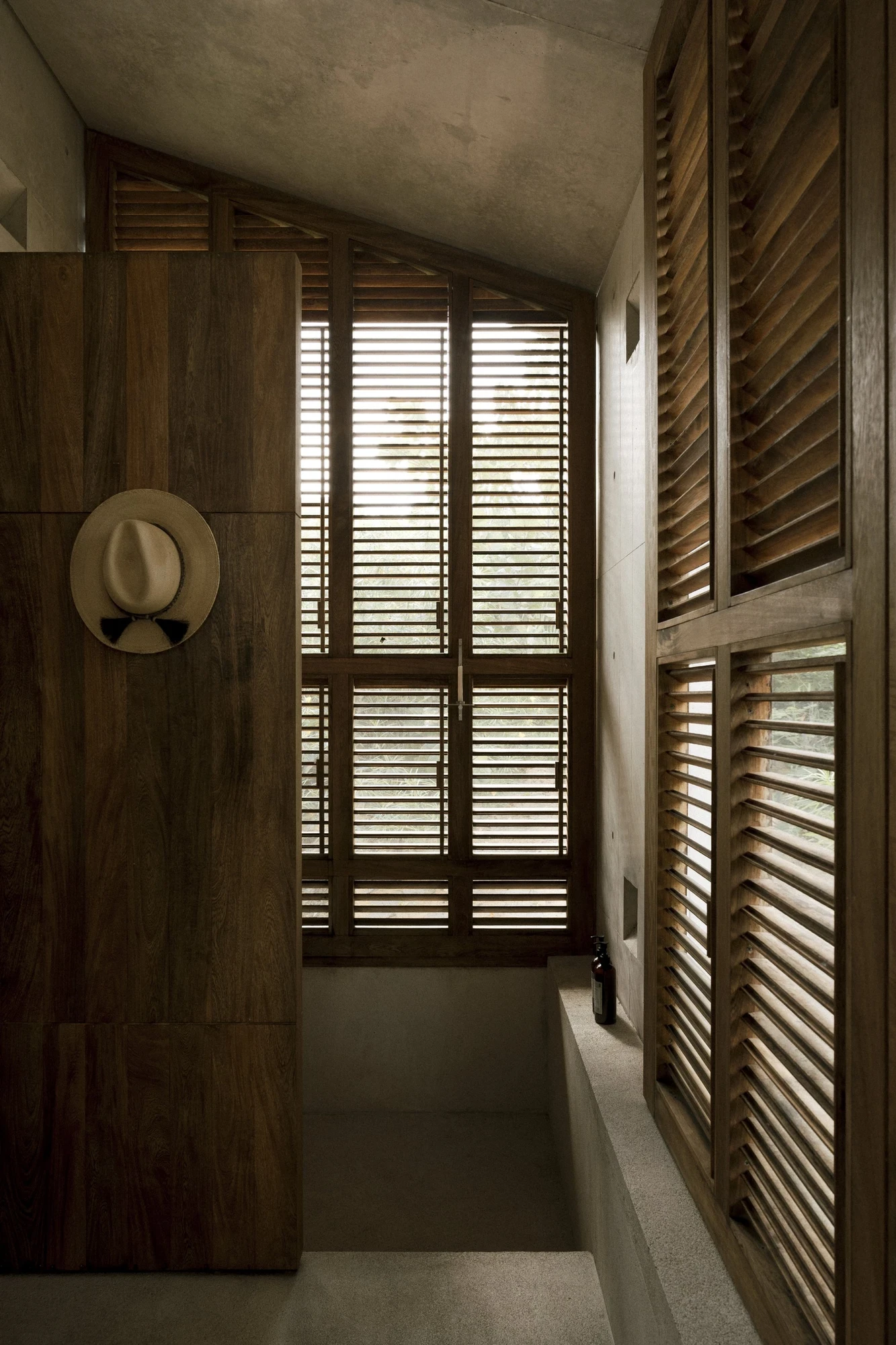 Casa Viento by Aranza De Ariño, photographed by Diego Orvaños — a straw hat hangs on a timber panel beside tall louvered shutters under a sloped concrete ceiling.