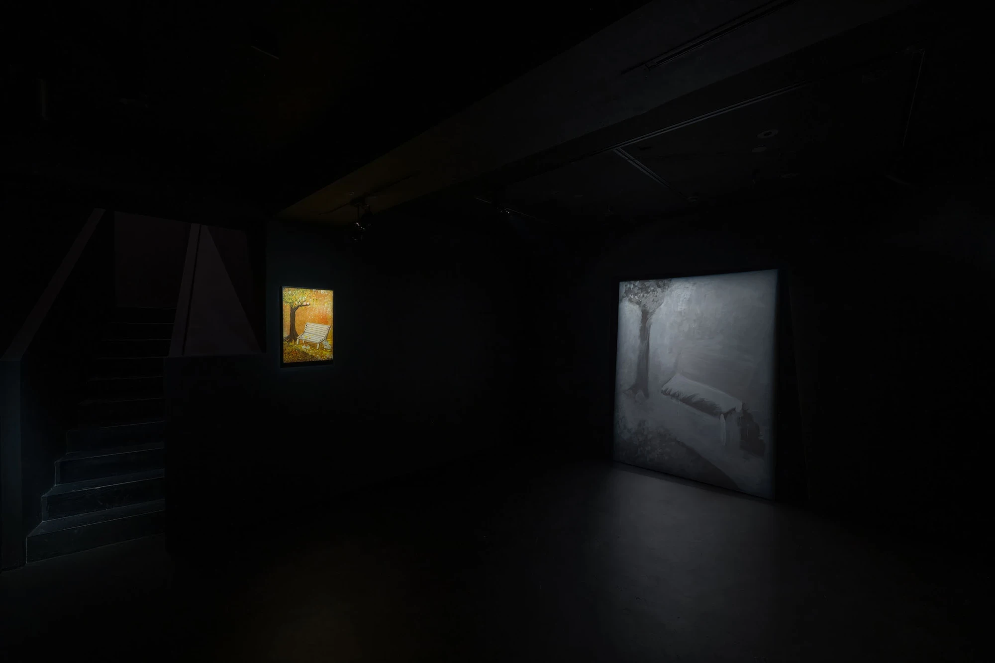 Spillover by Ann Zhao, photographed by Felix S.C. Wong — Installation view with the small ochre childhood painting lit on the left wall and the large grey monochrome canvas leaning at right in the dark room