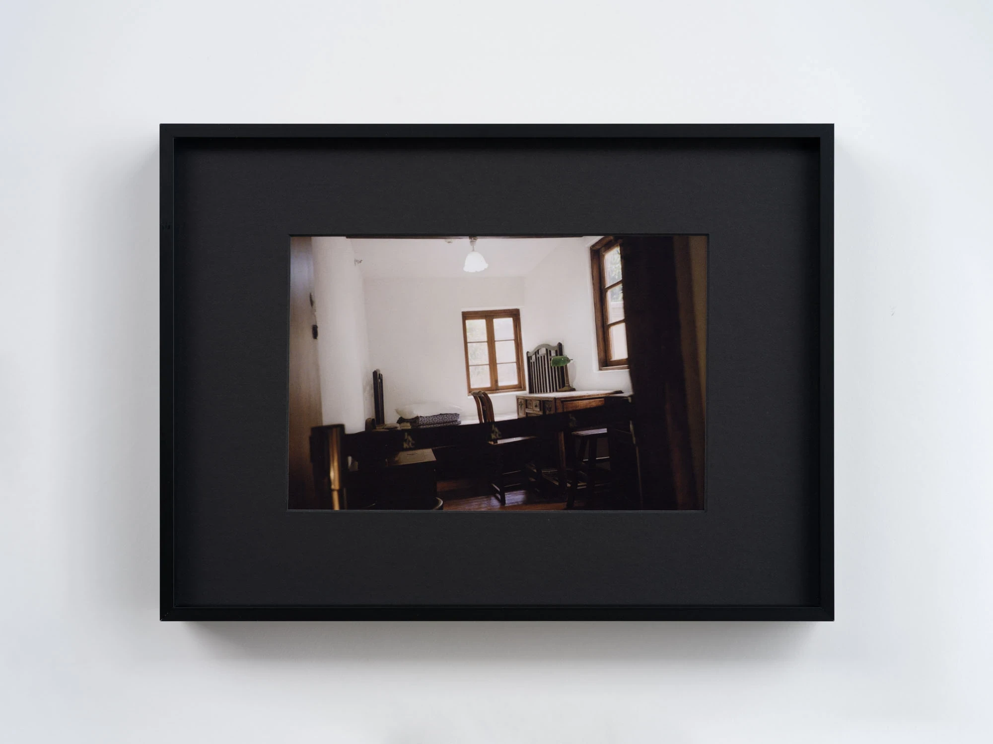 Spillover by Ann Zhao, photographed by Felix S.C. Wong — A framed colour photograph of a bare interior with two windows, dark wooden furniture and a green banker's lamp against white walls