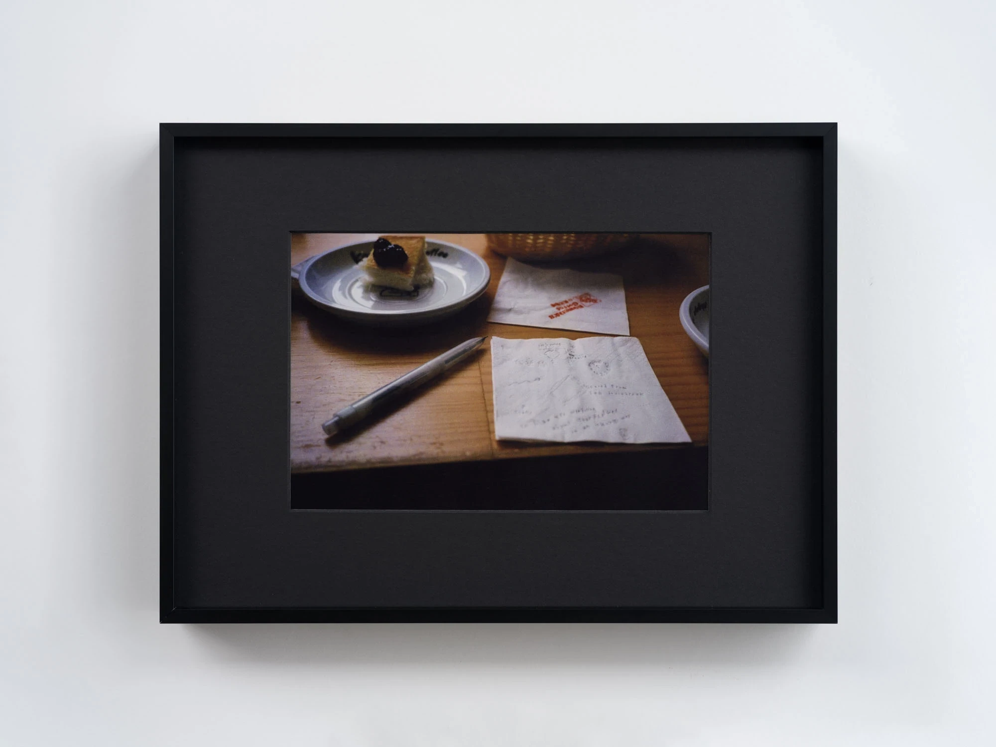 Spillover by Ann Zhao, photographed by Felix S.C. Wong — A colour photograph of a wooden cafe table with a slice of cheesecake, a pen and a napkin covered in faint handwritten notes