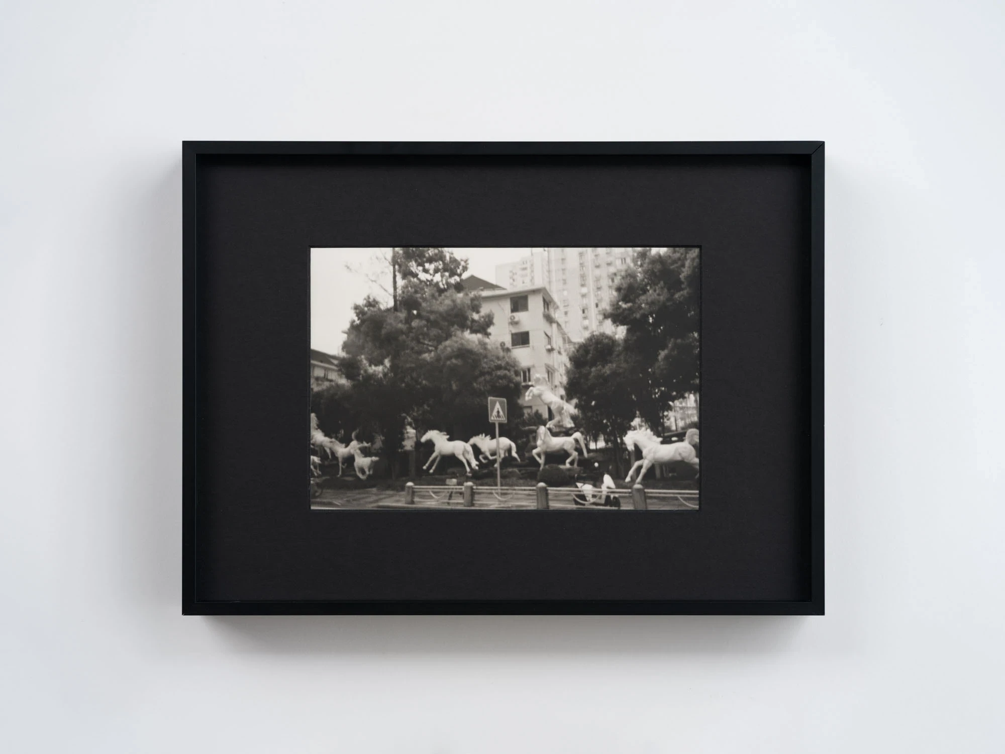 Spillover by Ann Zhao, photographed by Felix S.C. Wong — A black-and-white photograph of white sculptural horses caught mid-gallop in a planted courtyard beneath apartment towers