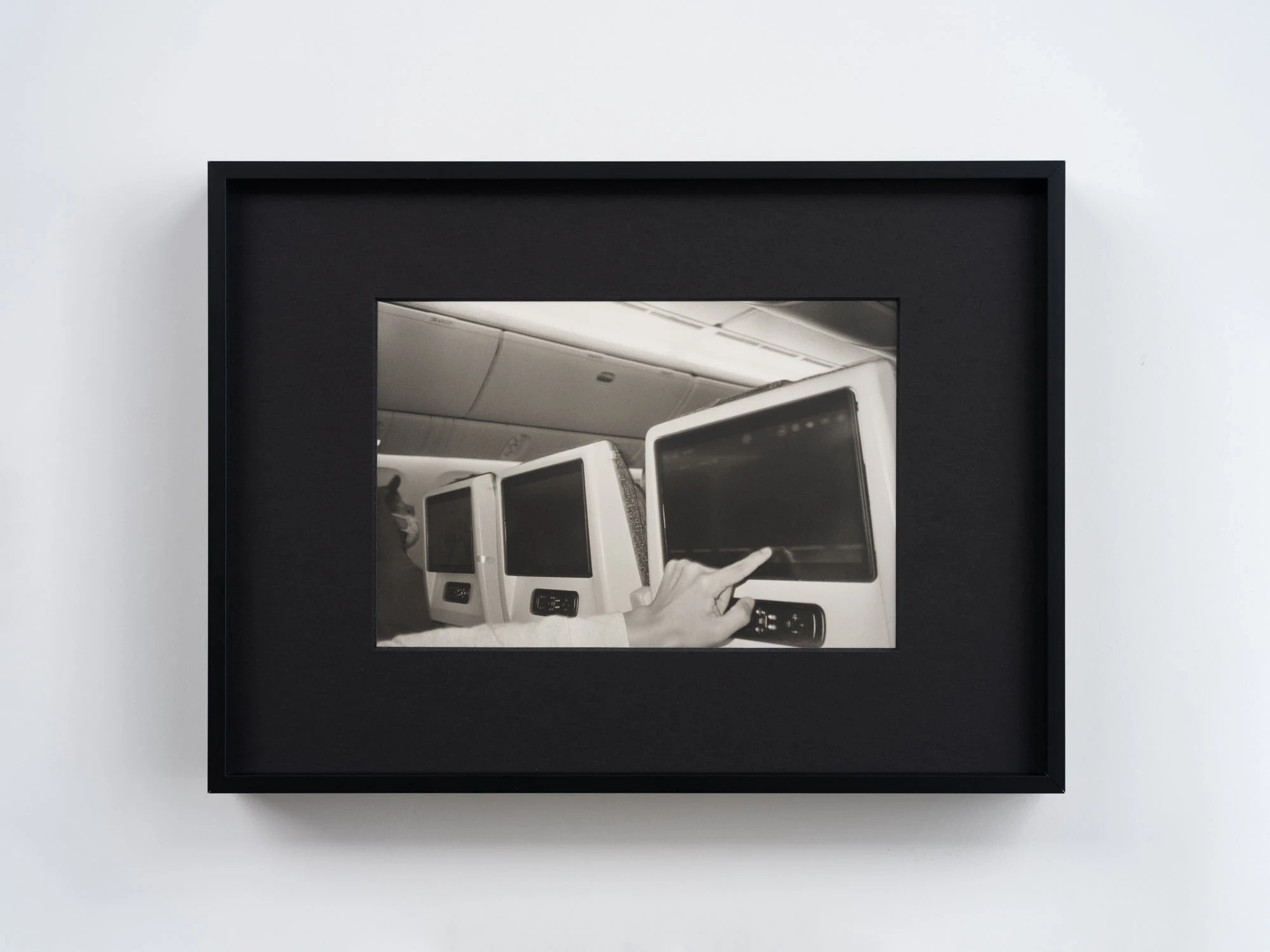 Spillover by Ann Zhao, photographed by Felix S.C. Wong — A black-and-white photograph of a hand reaching to touch an aircraft seatback screen, a row of dark monitors receding beside it