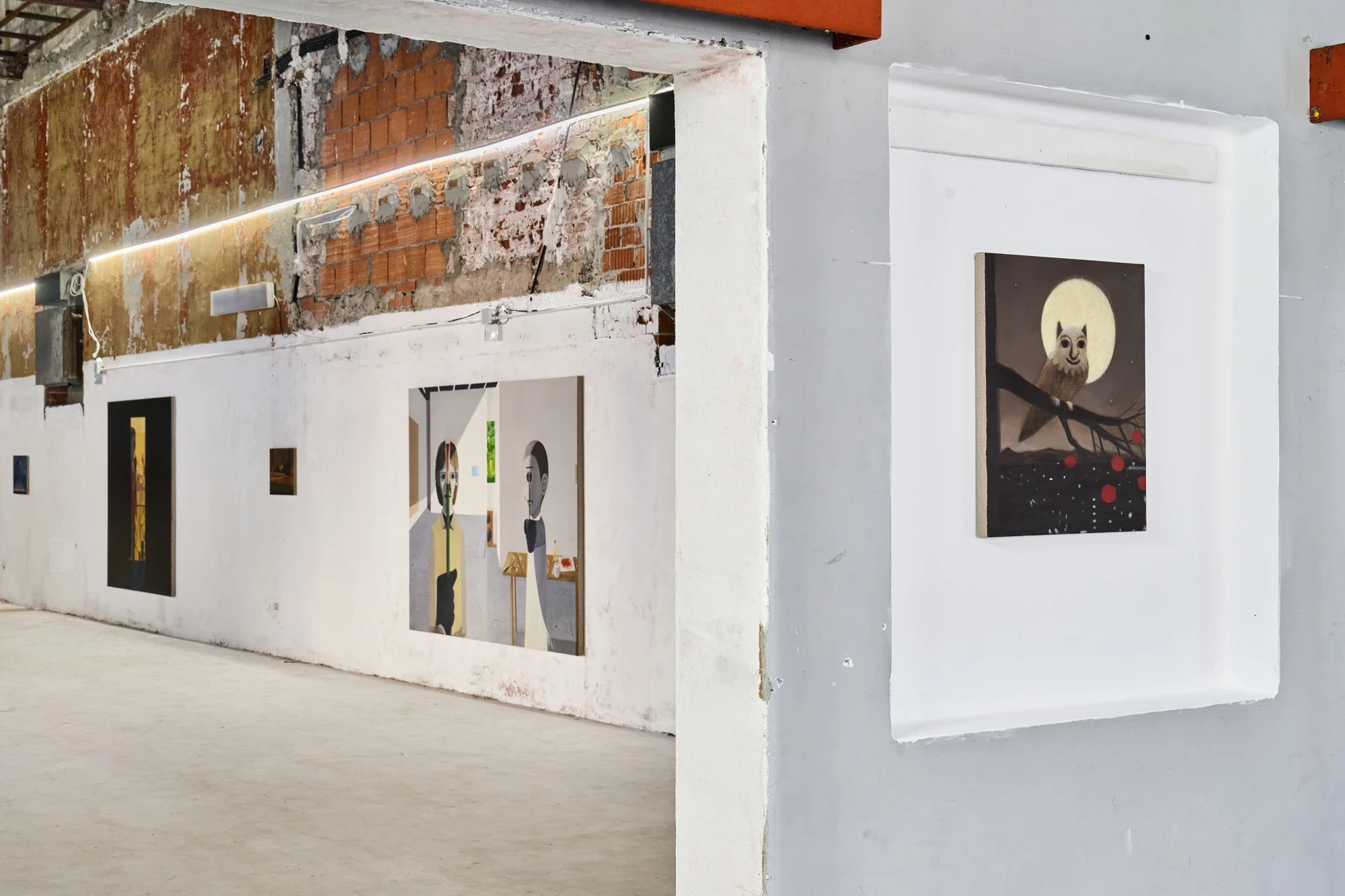 Every Day, Mostly Night, Sometimes Daylight by Satoru Kurata, photographed by Michela Pedranti -- A row of small dark paintings runs along a wall of exposed brick and ochre-stained plaster, with the owl-and-moon canvas set apart in a white cut-out niche o