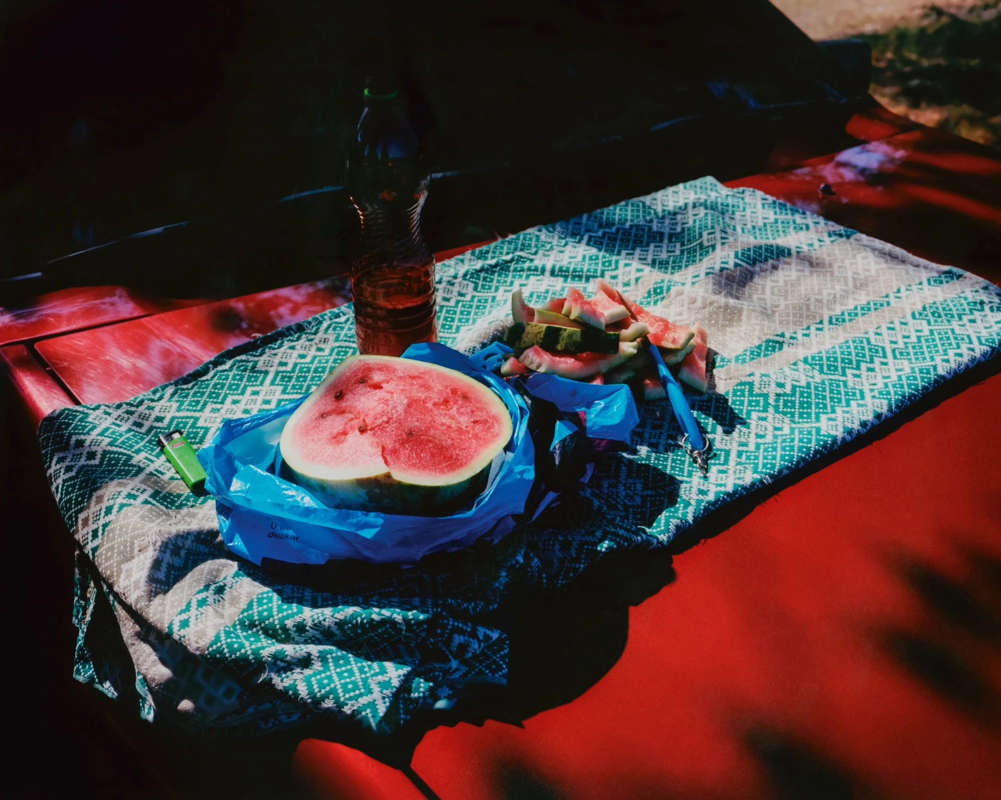 Unofficial Dnipro by Eugenia Skvarska and Kalpesh Lathigra — A halved watermelon and a bottle on an embroidered cloth spread over a red car bonnet.
