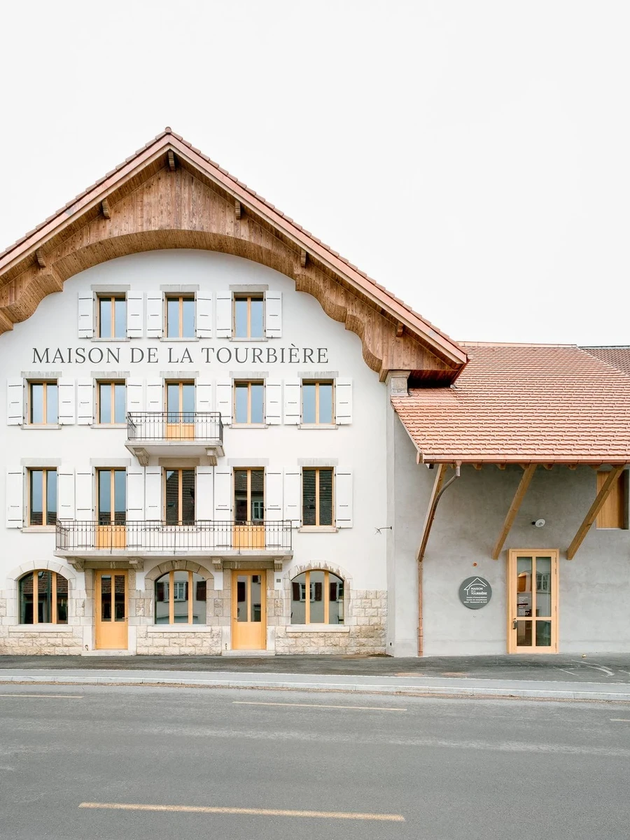 Maison de la Tourbiere by Frundgallina — Les Ponts-de-Martel, Switzerland: a pale rendered facade under a carved timber gable, "MAISON DE LA TOURBIERE" lettered across the render above a sandstone base.