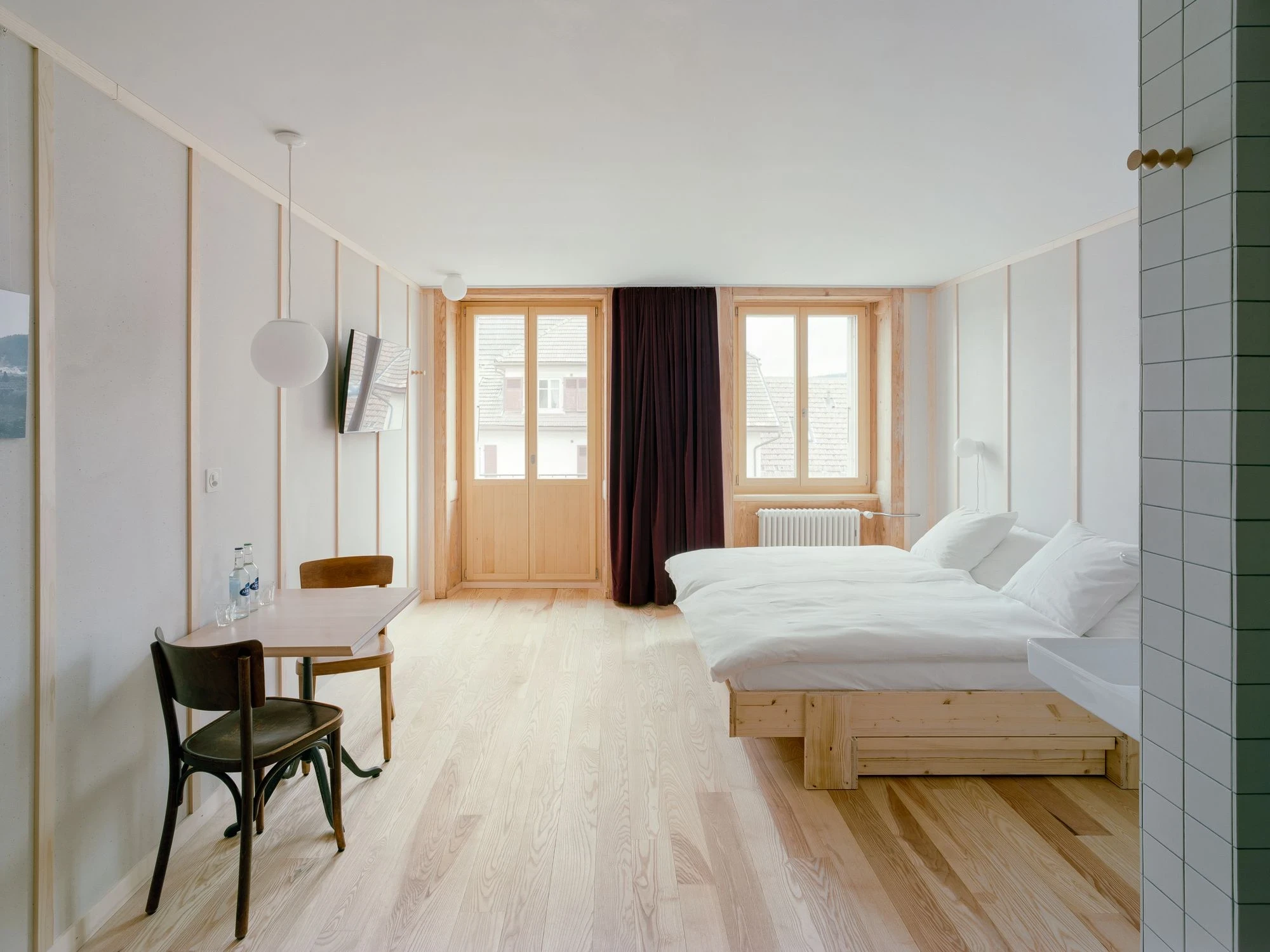 Maison de la Tourbiere by Frundgallina, photographed by Olivier Di Giambattista — A hotel room in pale ash boards and battened walls, a timber-framed door and window, dark curtain and a low solid-timber bed under a glass globe pendant.