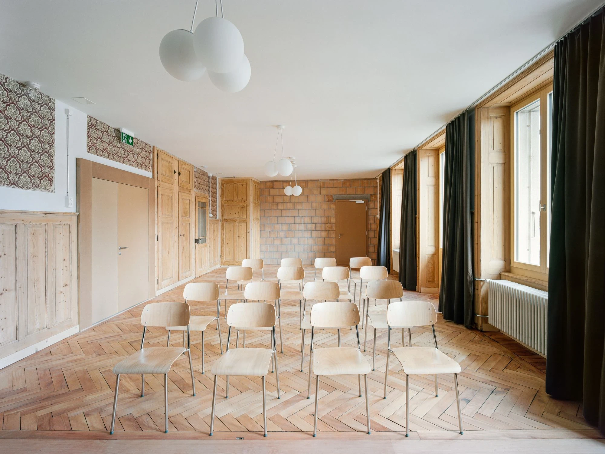 Maison de la Tourbiere by Frundgallina, photographed by Olivier Di Giambattista — The multipurpose hall with herringbone parquet, rows of pale plywood chairs, salvaged floral wallpaper, an exposed brick wall and olive velvet at the windows.