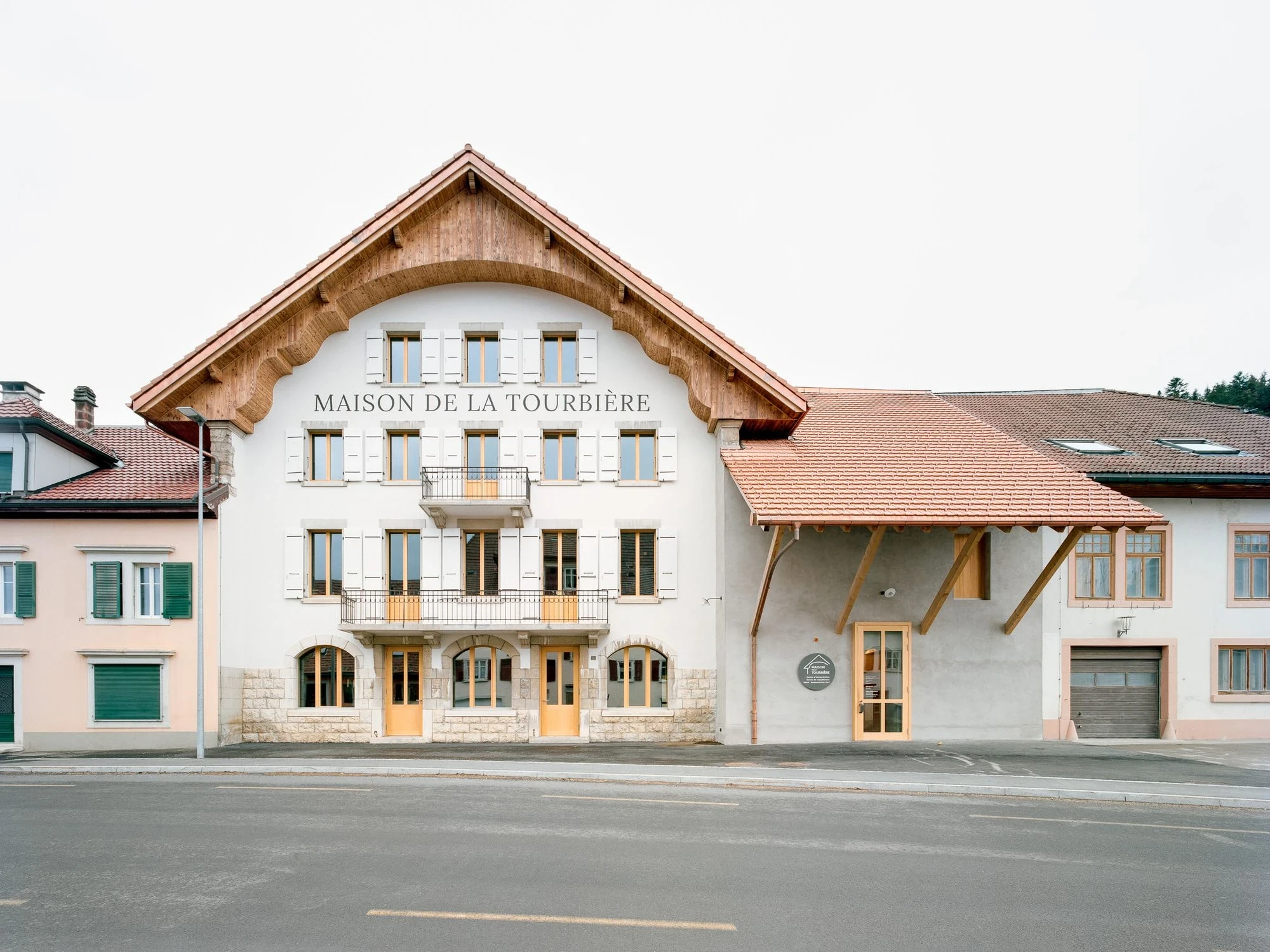Maison de la Tourbiere by Frundgallina — Les Ponts-de-Martel, Switzerland: a pale rendered facade under a carved timber gable, "MAISON DE LA TOURBIERE" lettered across the render above a sandstone base.