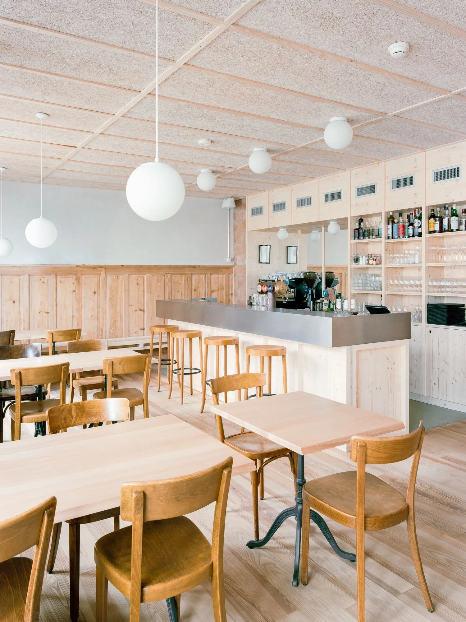 Maison de la Tourbiere by Frundgallina, photographed by Olivier Di Giambattista — The cafe seen from the bar, a stainless counter, pale ash tables and salvaged bentwood chairs under white glass globe pendants and a wood-fibre ceiling.