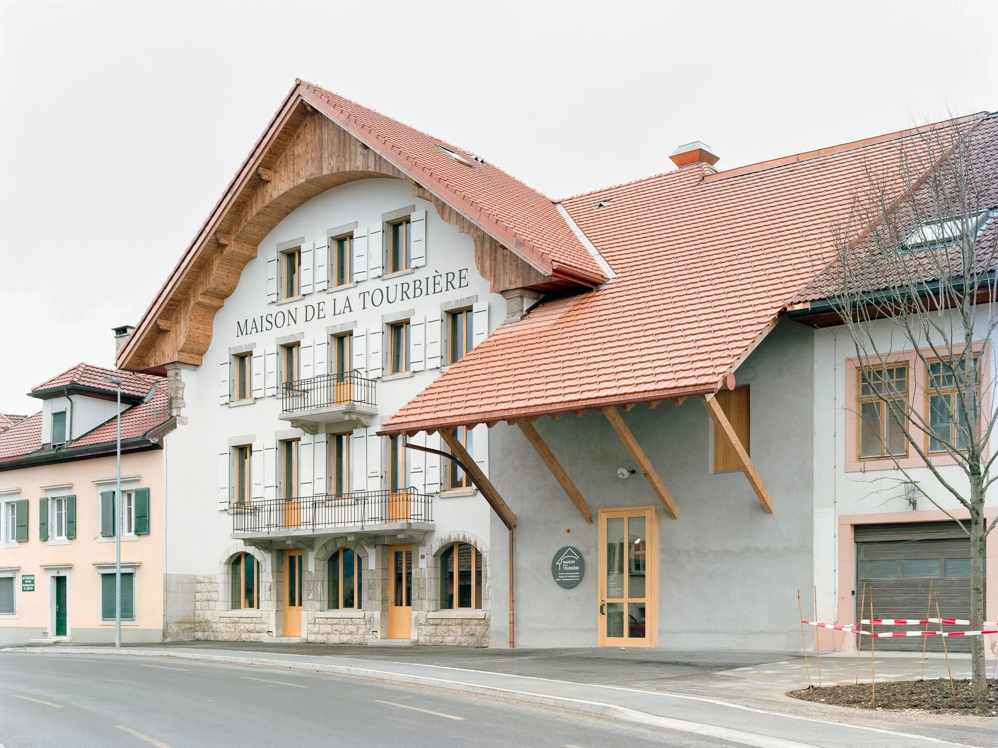 Maison de la Tourbiere by Frundgallina, photographed by Olivier Di Giambattista — The building seen at an angle, the tiled entrance canopy on timber brackets projecting over a rendered side wall beside the pale gabled front.