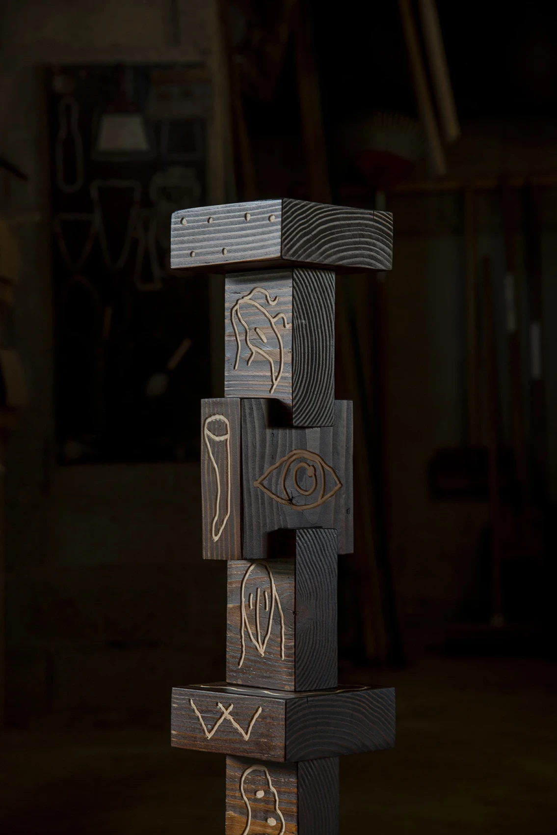 Les Rhizopodes by Barreau Charbonnet — A vertical totem of shifting timber blocks, each face carved with an eye, a seed and a single-cell form picked out in cream