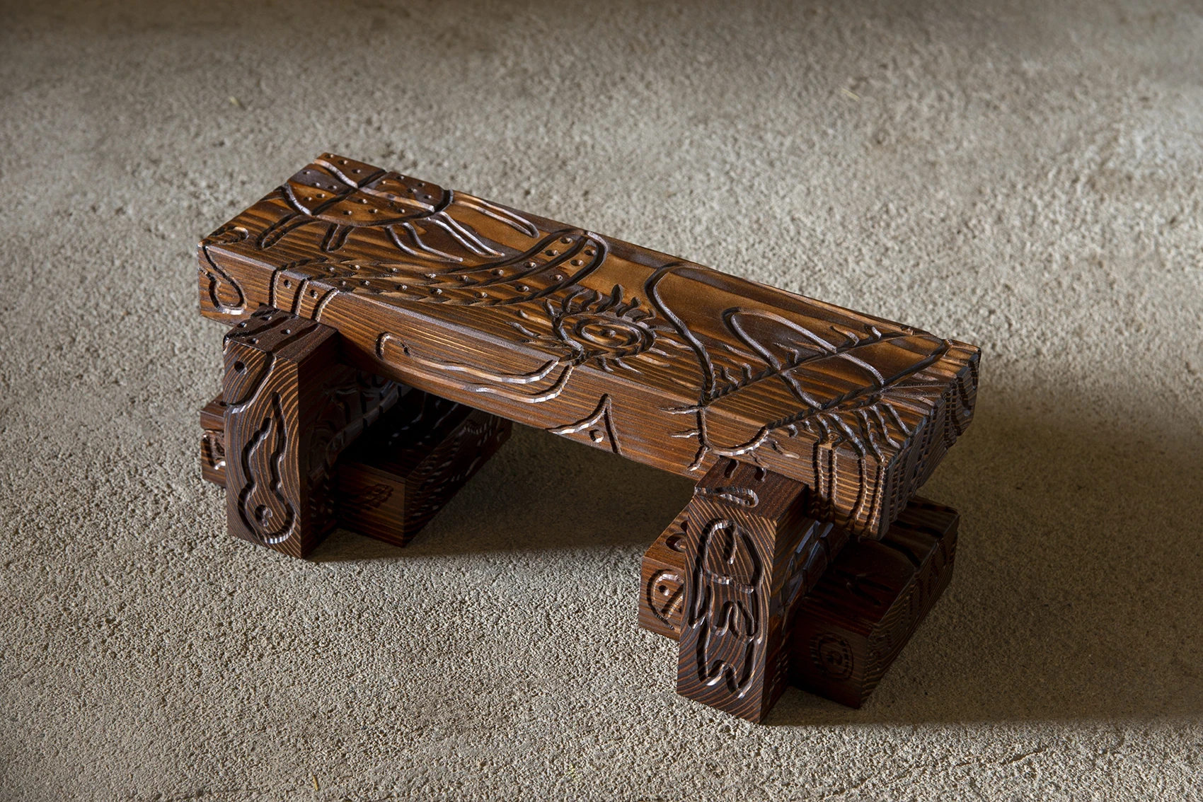 Les Rhizopodes by Barreau Charbonnet — A small bench at three-quarters, spider and sun motifs carved into the natural, unpainted timber