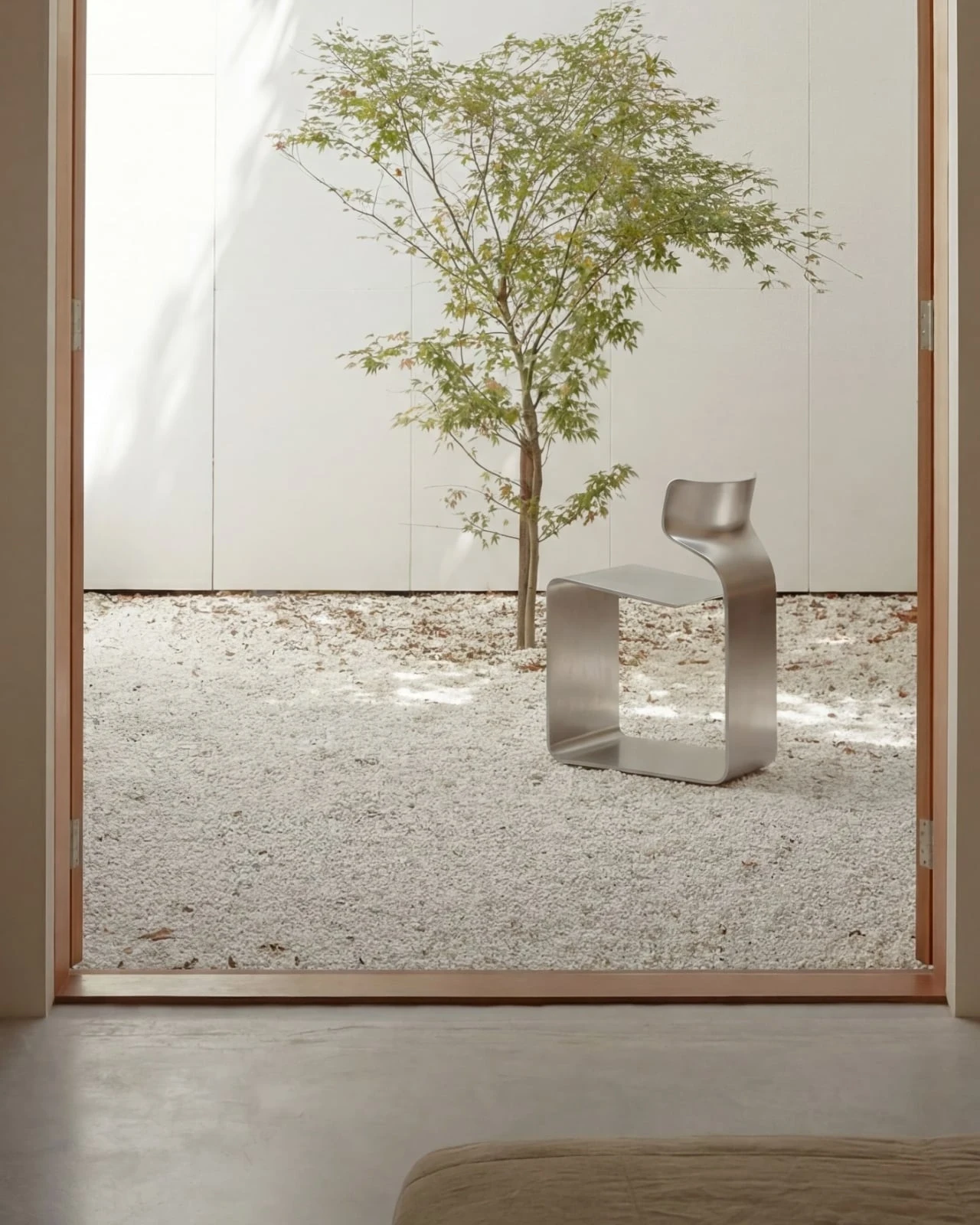 ZERO Chair by Davide Bozzo — The chair seen through a doorway on a bed of pale gravel beside a young maple, its brushed surface catching the daylight of the courtyard