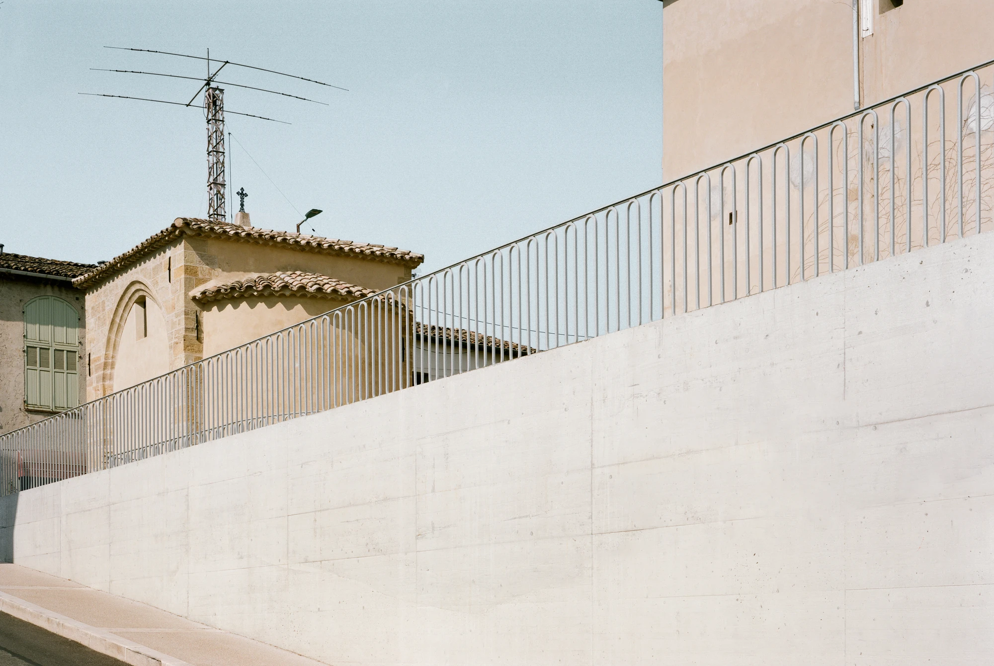 Biot Municipal Building Extension by Atelier EGR, photographed by Giaime Meloni — A curving concrete retaining wall topped by looped balustrades, with the chapel roof and village rooftops behind.