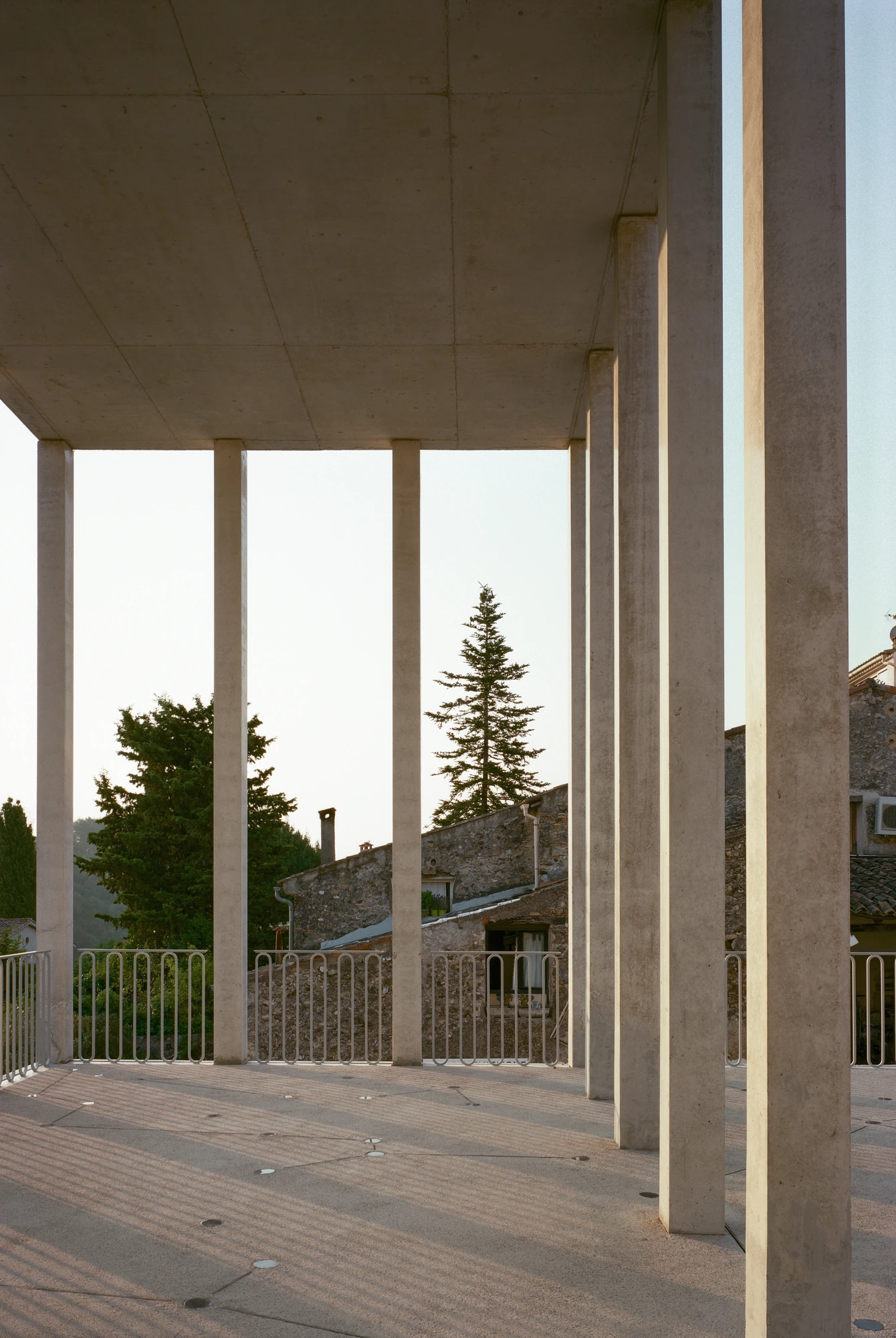 Biot Municipal Building Extension by Atelier EGR, photographed by Giaime Meloni — View out from under the upper level, slim concrete columns and looped metal balustrades framing the village roofs and trees.