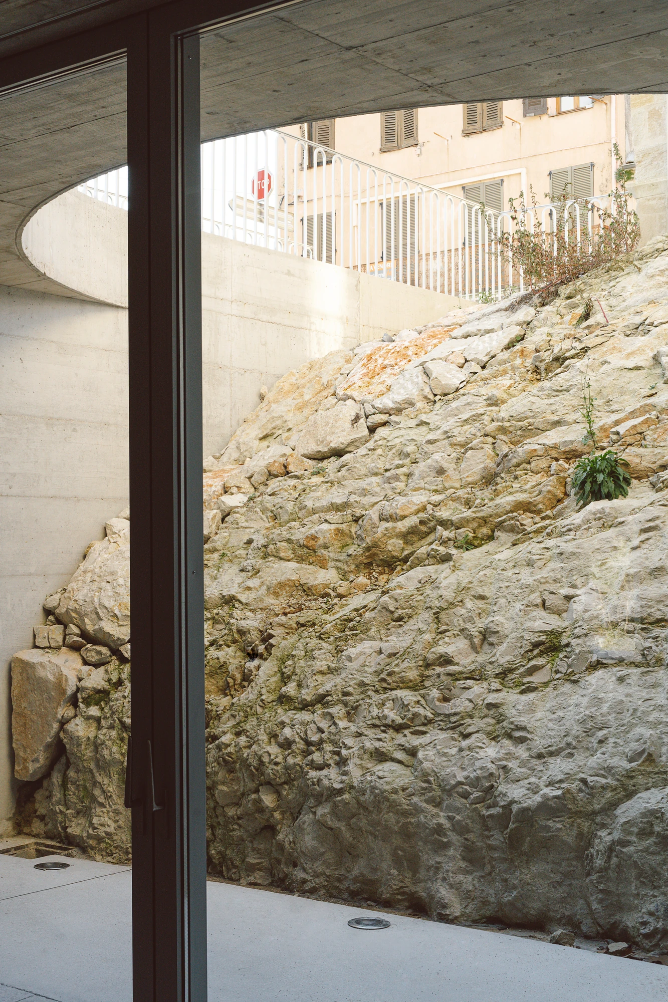 Biot Municipal Building Extension by Atelier EGR, photographed by Giaime Meloni — Through the glazing, the excavated limestone of the rear patio rising toward a village house at street level.