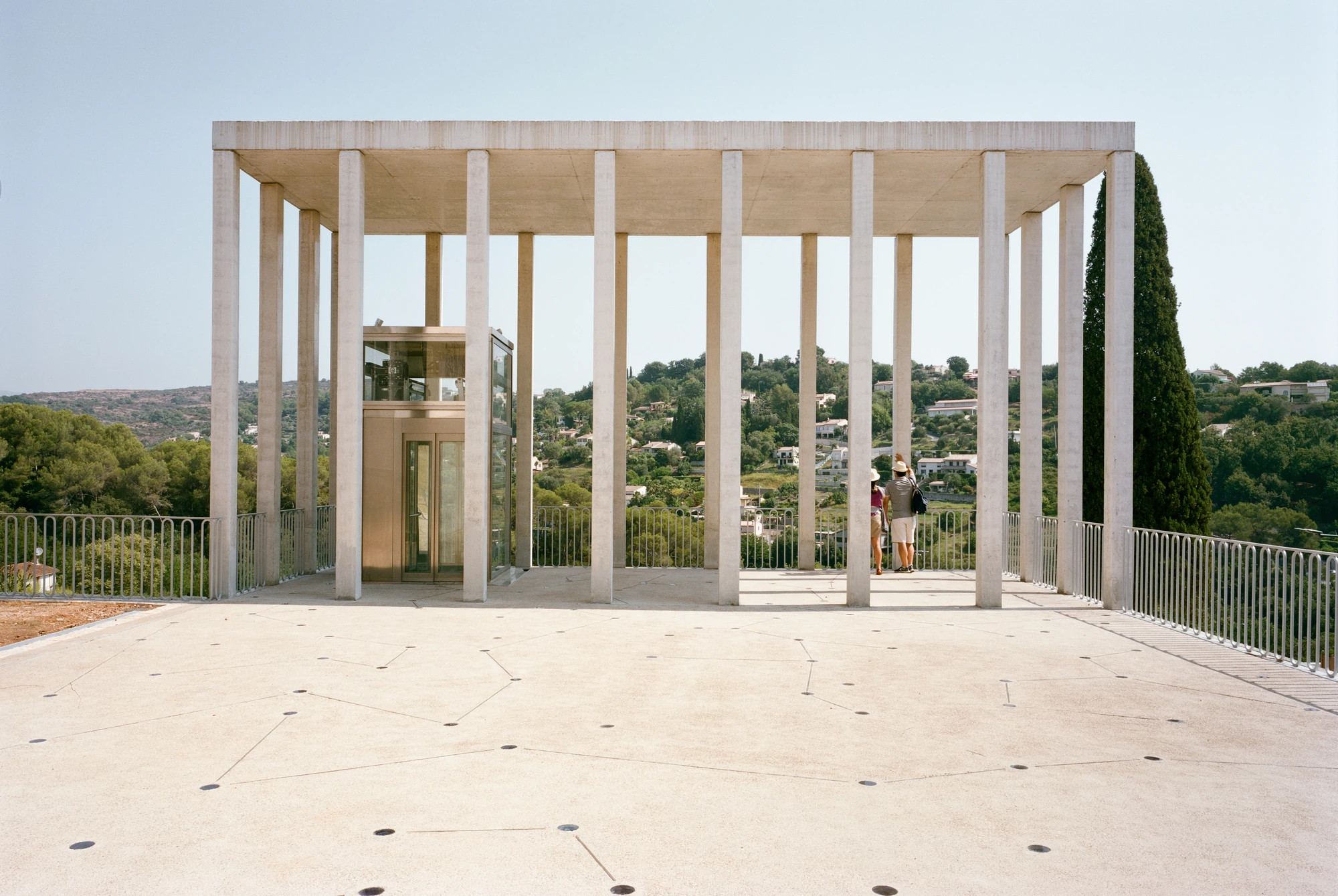 Biot Municipal Building Extension by Atelier EGR, photographed by Giaime Meloni — The belvedere deck with the glazed lift standing among the columns and two people looking out over the hills.