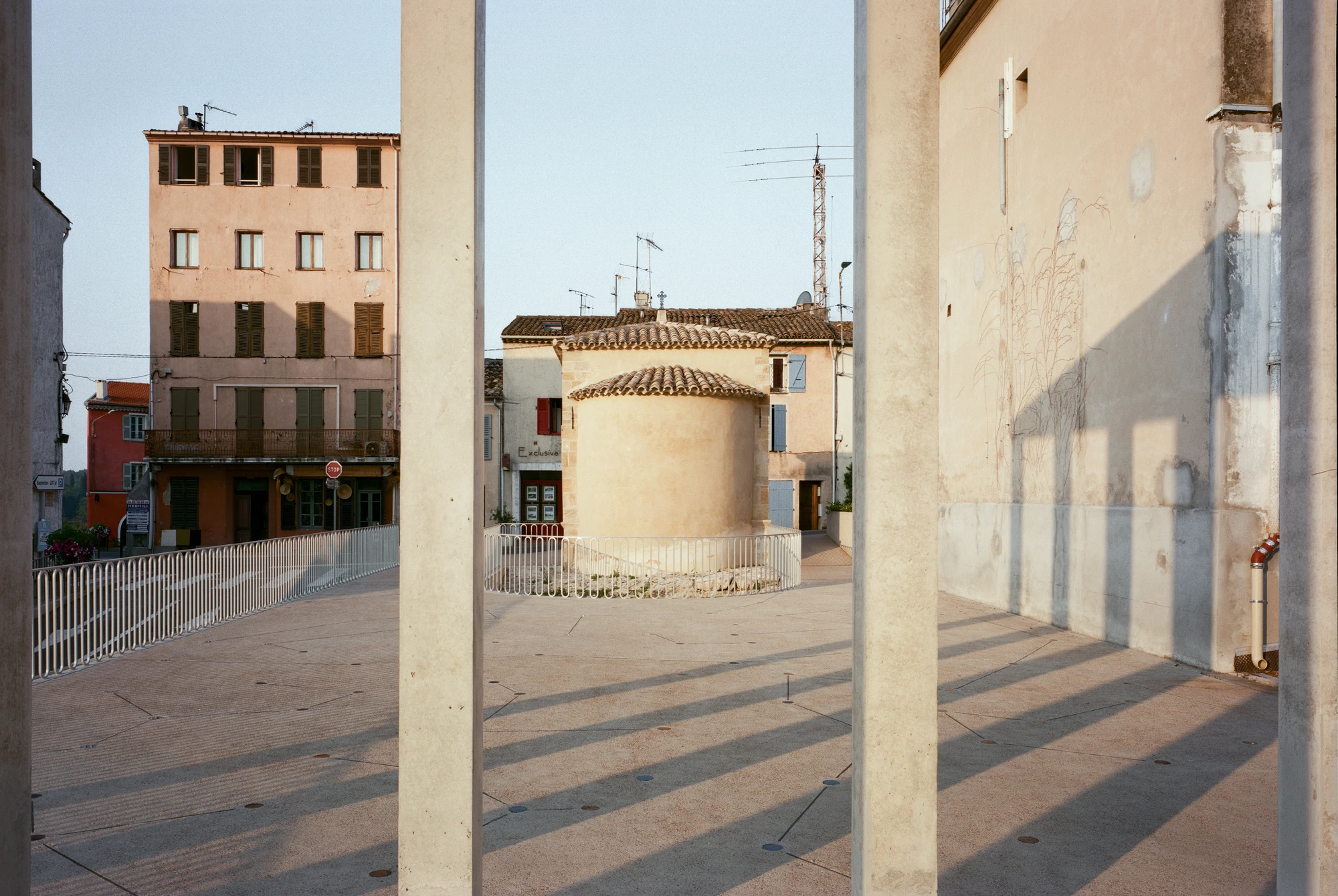 Biot Municipal Building Extension by Atelier EGR, photographed by Giaime Meloni — The public square seen between two columns, long shadows crossing the paving toward the rounded apse of the chapel.