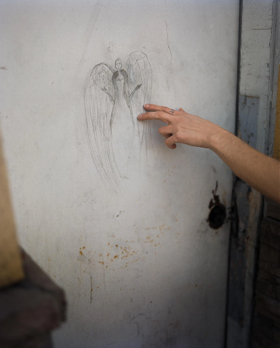 King, Queen, Knave by Gregory Halpern, photographed by Gregory Halpern — A bare arm reaches in from the right, fingertips resting on a pencil drawing of a winged figure on a white painted door beside a rusted lock.