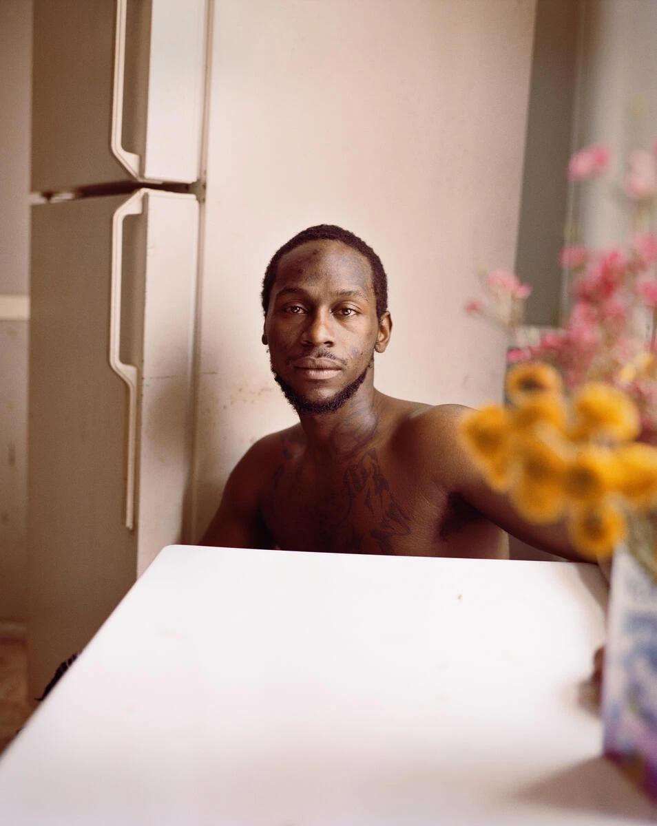 King, Queen, Knave by Gregory Halpern, photographed by Gregory Halpern — A shirtless man with chest tattoos sits at a white table beside a vase of yellow and pink flowers, a refrigerator at his shoulder.