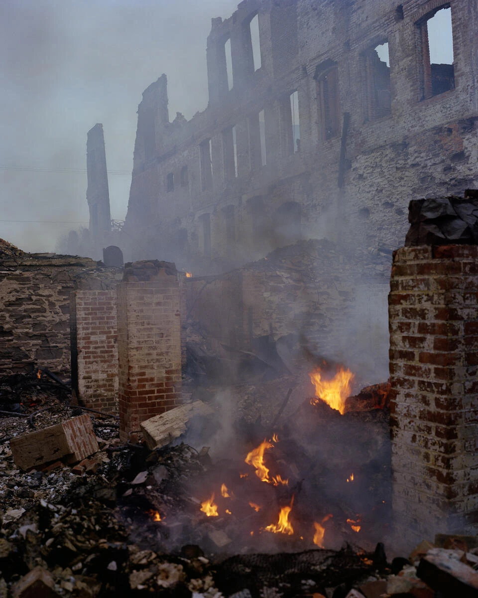 King, Queen, Knave by Gregory Halpern, photographed by Gregory Halpern — Flames burn through rubble between brick piers, smoke rising against the gutted upper floors of a stone and brick building.