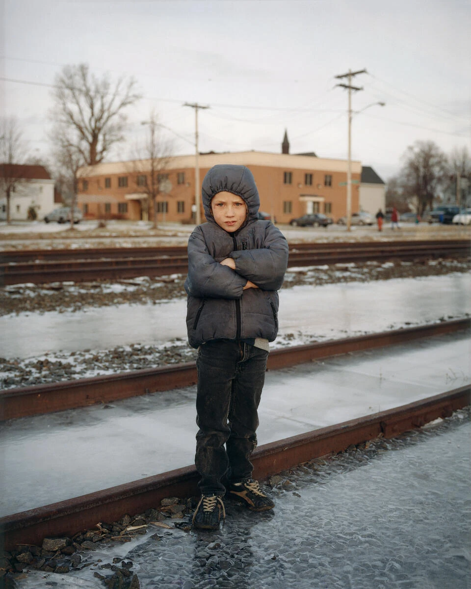 King, Queen, Knave by Gregory Halpern, photographed by Gregory Halpern — A boy in a grey hooded coat stands with his arms crossed on a railway line, ice between the rails and a low brick building behind him.